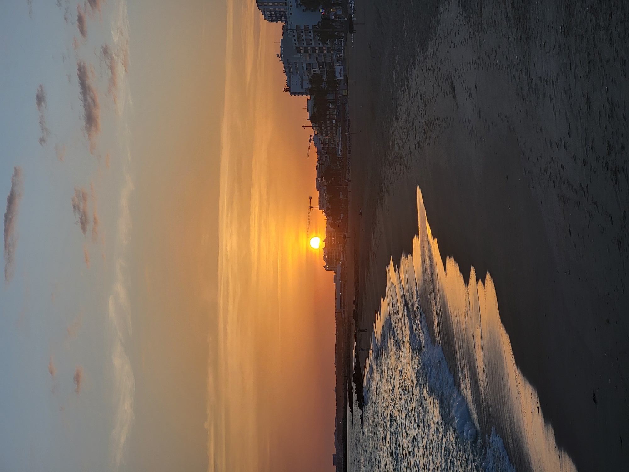 The image depicts a sunset on a beach in Quarteira, Portugal. The sun, a bright orange orb, is low on the horizon, casting a warm glow across the sky and reflecting on the wet sand. The ocean is calm with gentle waves lapping at the shore. In the background, buildings line the coast, with palm trees scattered between them. A few people are visible on the beach, appearing as small silhouettes against the sand. The scene conveys a peaceful and serene atmosphere.