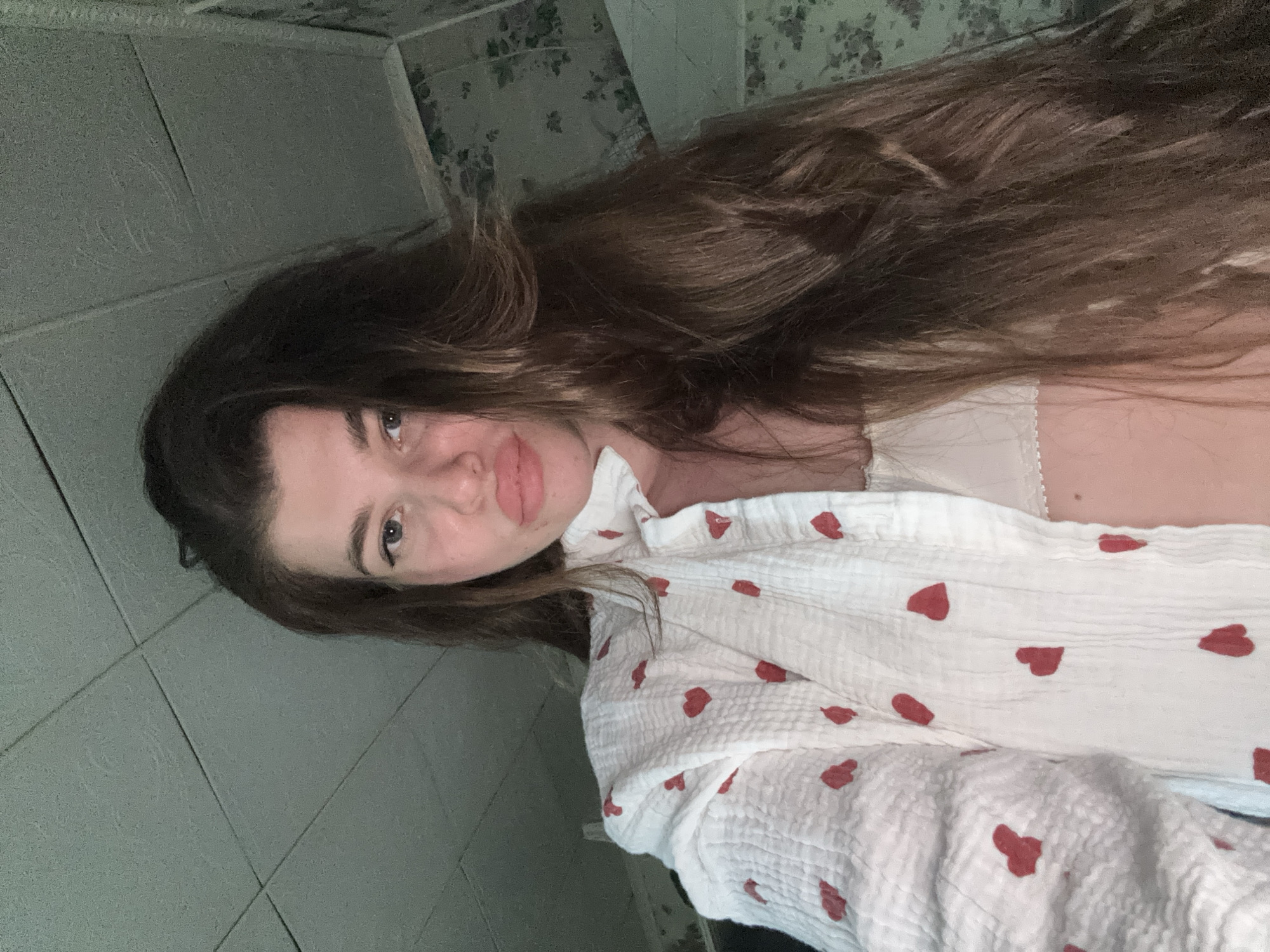 A selfie shot shows a woman with long, brown hair wearing a white top with red heart patterns. The background appears to be a bathroom with light green tiles and a floral wallpaper border near the ceiling. There is no verifiable evidence to suggest the location is in the city of Мещёры, Russia.