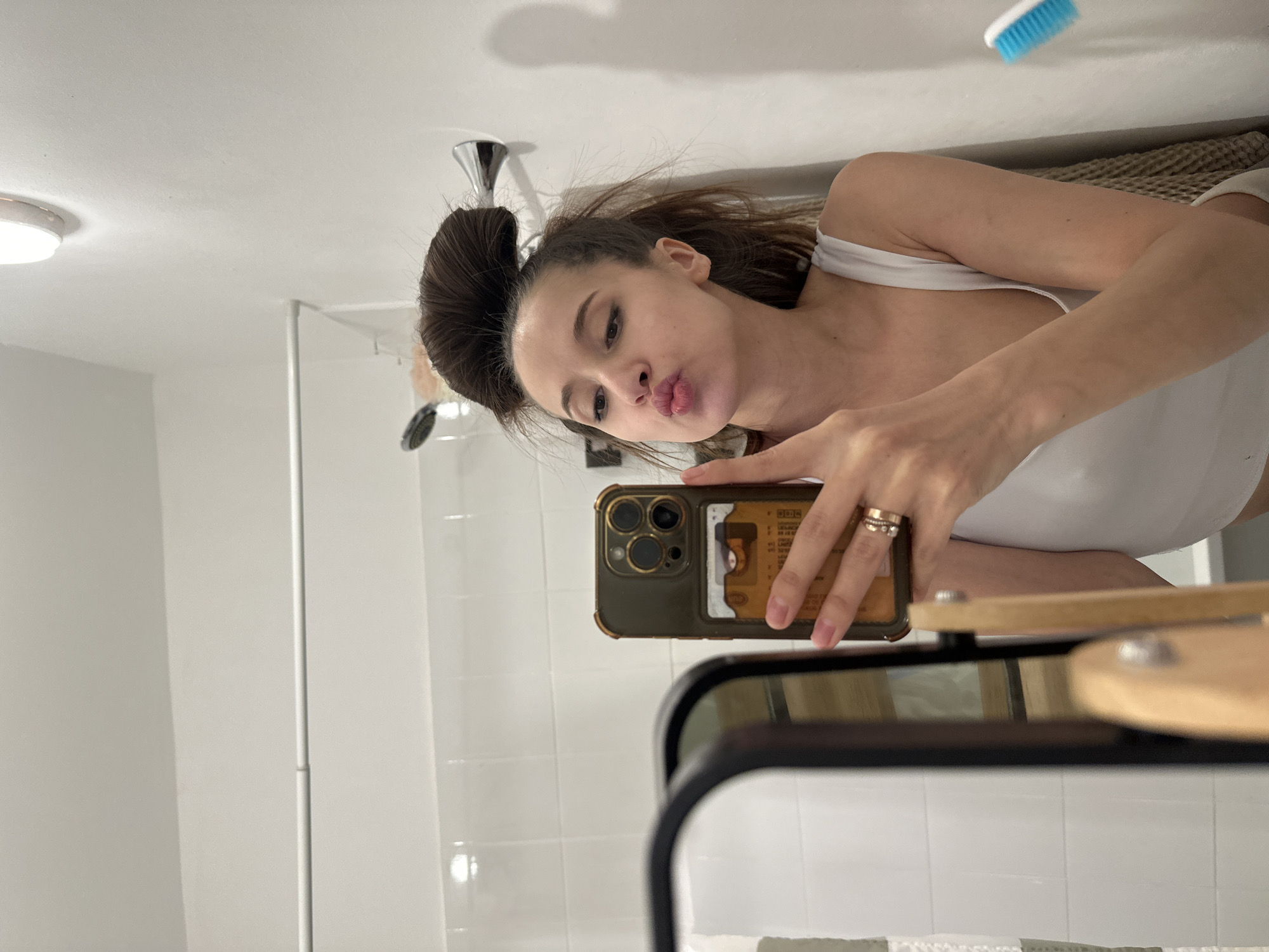 The image is a mirror selfie featuring a young woman in a modern bathroom setting. The woman, appearing to be in her 20s or early 30s, is positioned slightly to her right, wearing a white tank top or camisole. Her dark hair is styled in a high bun, with some loose strands around her face. She is making a playful "pouty" or "duck" face towards the camera, conveying a casual and self-confident mood.
She holds a dark olive-green or brown smartphone, likely an iPhone Pro model, in her right hand, which is adorned with multiple rings on her ring finger. A transparent card holder is attached to the back of the phone case, revealing several indistinct cards within. The phone partially obscures her lower face and chest.
The background indicates a clean, white-tiled bathroom. A chrome shower head is visible to the left of her head. A bright, round ceiling light fixture is seen in the upper left, illuminating the space. In the upper right corner, a small blue and white brush is visible. The dark frame of the mirror is prominent in the lower right, reflecting a light-colored wooden shelf or countertop beneath it. The overall environment is simple and functional. No specific text is legible on the cards in the phone case or elsewhere. The location context provided is Yekaterinburg, Russia, though no specific visual cues within the image confirm this.
