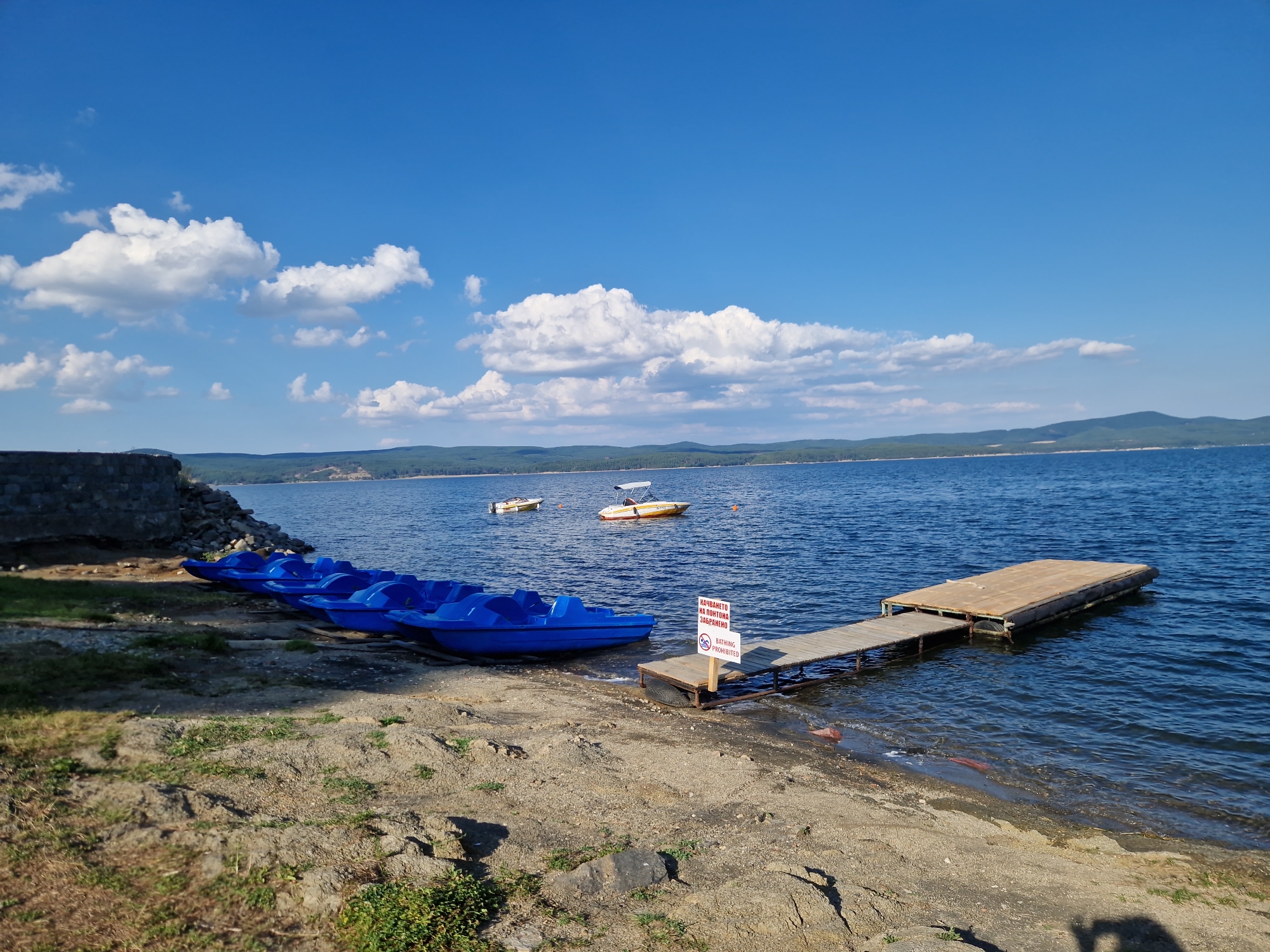 The image captures a serene lakeside scene on a sunny day in Dolni Pasarel, Bulgaria. A clear blue sky dotted with fluffy white clouds stretches above a vast expanse of deep blue water. In the foreground, a sandy and rocky shoreline leads to the water's edge. Several bright blue pedal boats are lined up on the shore, suggesting a recreational area.
To the right, a wooden pier extends into the lake, with a flat platform at its end. A sign attached to the pier reads "BATHING PROHIBITED" in both Bulgarian and English, indicating a restriction on swimming in this area. In the middle ground, two small motorboats are anchored in the water. The distant shoreline is lined with lush green trees and rolling hills, creating a picturesque backdrop. The overall atmosphere is peaceful and tranquil, evoking a sense of a perfect summer day by the water.