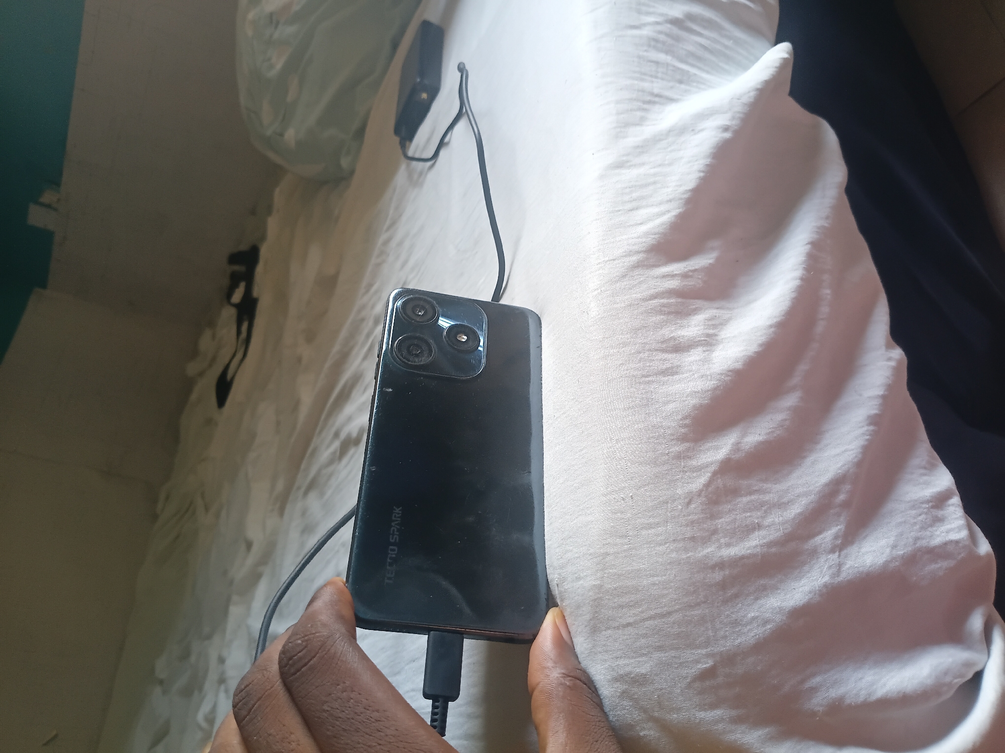 A close-up shot focuses on a black Tecno Spark smartphone lying on a white bedsheet. The phone is connected to a charging cable, which snakes across the bed towards a black power adapter in the background. A person's hand, with dark skin, is partially visible on the left side of the frame, holding the phone. The environment appears to be a bedroom in Jalingo, Nigeria, with a blurred background suggesting other bedding and furniture. The lighting indicates it's daytime, possibly indoors with natural light.