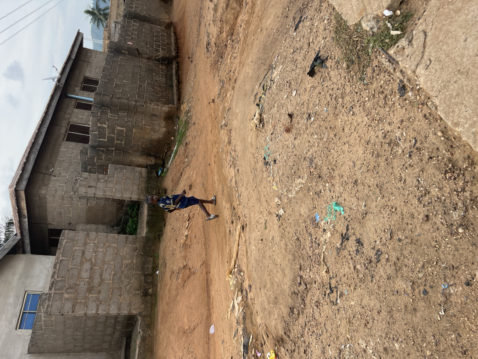 The image displays an outdoor scene featuring a young boy on a dirt path, flanked by buildings. The boy, positioned towards the left side of the frame, is wearing a blue short-sleeved shirt with yellow patterns, blue shorts, and light-colored flip-flops. He appears to be walking or standing on the uneven, light brown dirt ground, which is strewn with small pebbles and various pieces of litter, including plastic bags and torn paper.
On the left side of the path, several buildings constructed from unrendered concrete blocks are visible. One prominent structure has a corrugated metal roof, rectangular windows, and a satellite dish mounted on its roof. Further down, another building features a distinct blue-framed window, suggesting a different style or stage of construction. In the background, a palm tree stands out against a cloudy sky. To the right of the dirt path, a concrete curb or raised section of ground is visible, also with some sparse vegetation and debris. The scene is illuminated by bright natural daylight, indicating it is daytime. No specific activities beyond the boy's presence are depicted, and no text is visible within the image. The setting suggests a semi-urban or rural environment, possibly in a developing region.