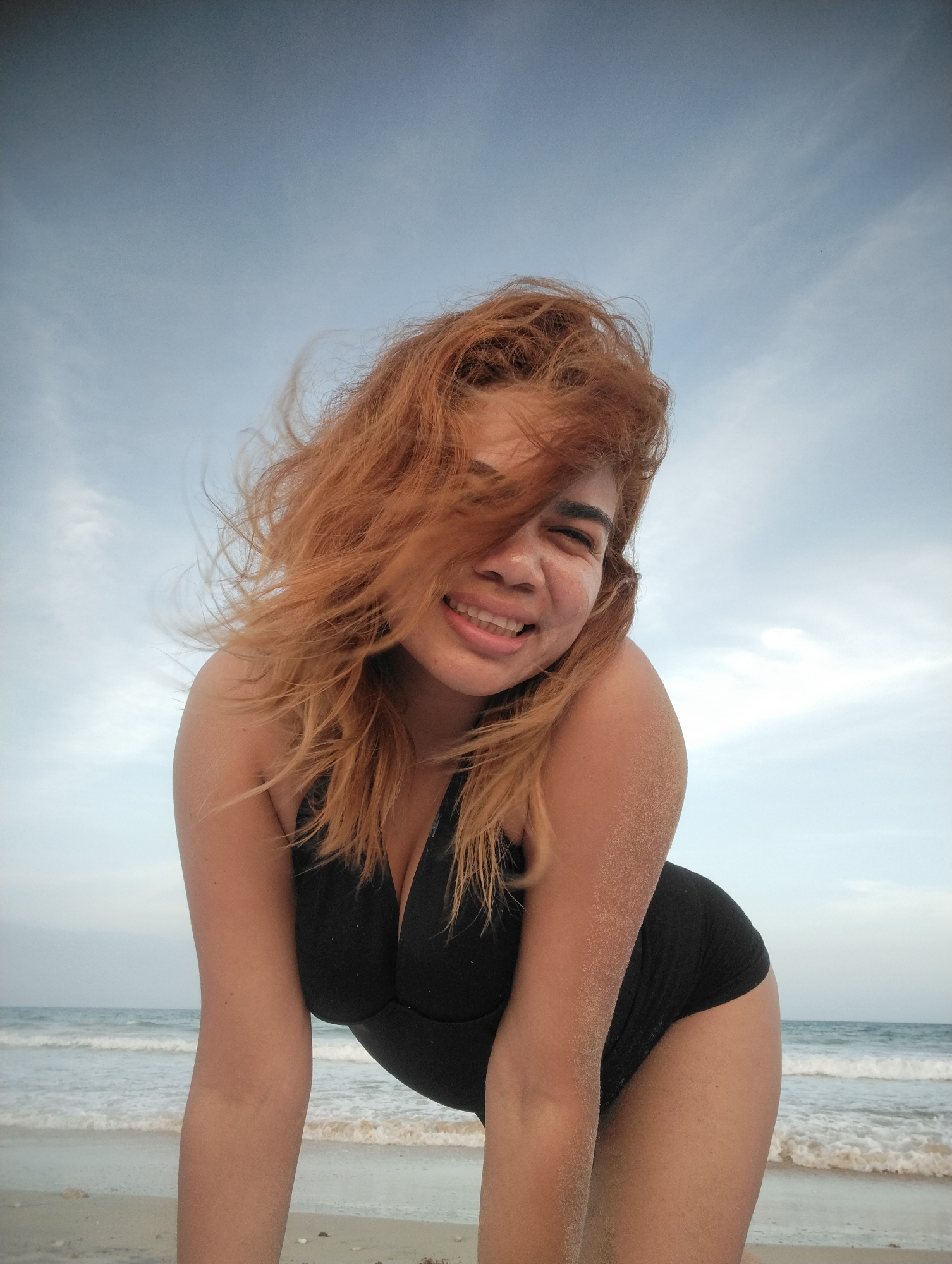 A woman with red-orange hair is at the beach, smiling at the camera. She's wearing a black one-piece swimsuit, and her skin is covered in sand. The ocean is visible in the background, with small waves coming ashore. The sky is a light blue with wispy clouds. While the prompt asks for the location as Porlamar, Venezuela, this cannot be verified from the image alone. The image only shows a woman, beach, and sky.