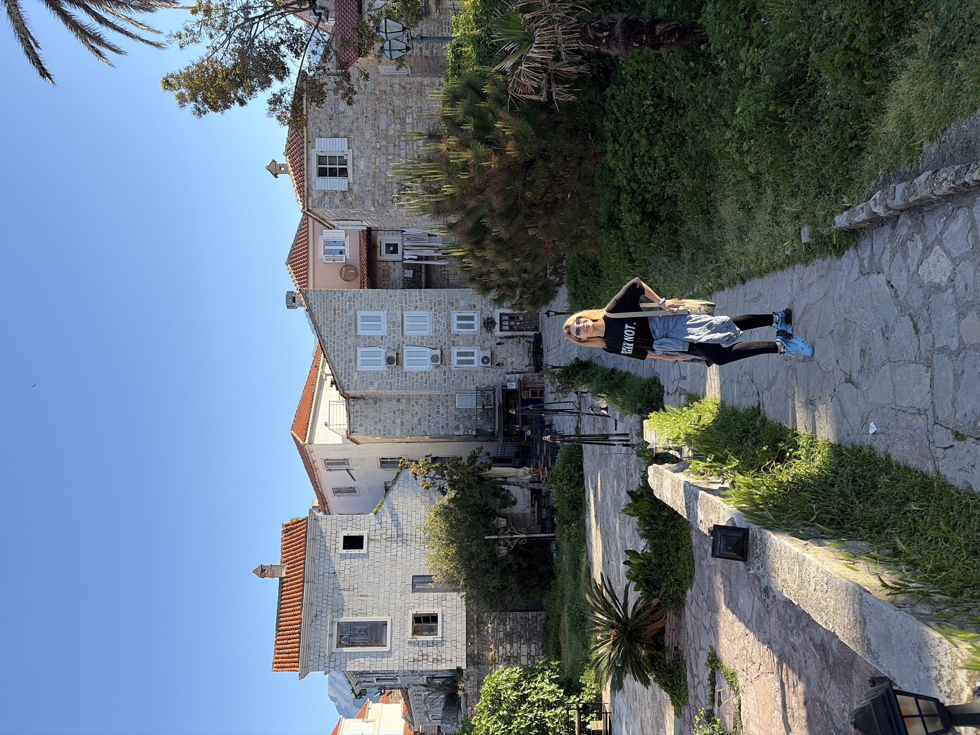 A woman stands on a stone path in the city of Budva, Montenegro. She wears a black t-shirt with white lettering, black leggings, and blue shoes, and carries a purse. Behind her are stone buildings with orange tiled roofs and white shutters. Lush green vegetation grows along the pathway and around the buildings. The sky is clear and blue.