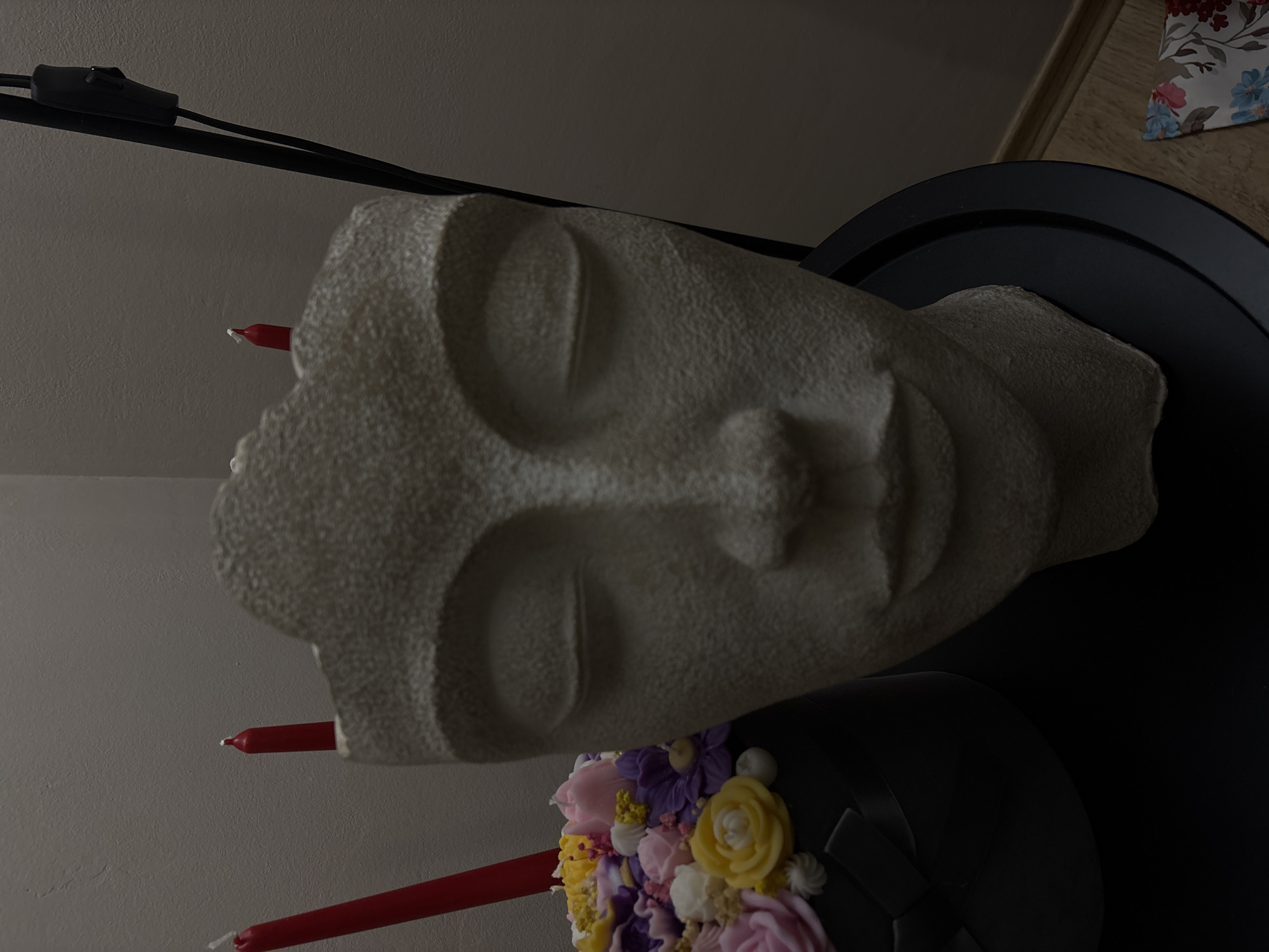 A textured white sculpture of a serene face with closed eyes is centered and rotated counter-clockwise. The face is mounted on a black tiered base decorated with colorful artificial flowers. Three red candles are inserted into the sculpture. A black electrical cord is visible in the top left. The background is a plain off-white wall. A sliver of light-colored wooden flooring is seen at the top right. There is no indication of the location being Veliko Tarnovo, Bulgaria.