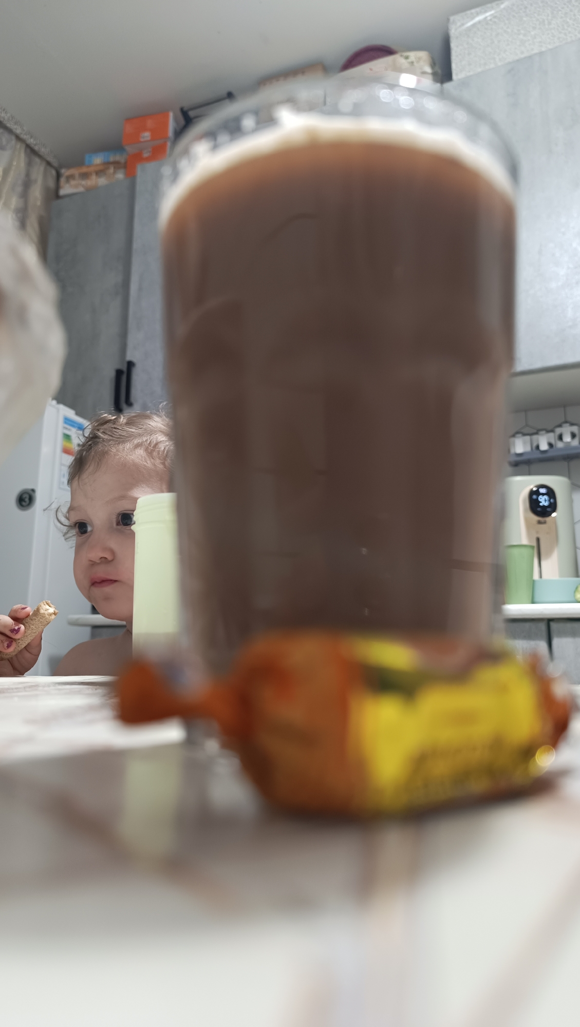 The image captures a close-up view of a child eating at a table in what appears to be a kitchen. The child, who has light-colored curly hair, is looking to their left while holding a piece of food in their right hand. In the foreground, a large, blurry glass of a brown beverage dominates the scene, with a blurred orange and yellow candy wrapper in front of it. Behind the child, a pale yellow cup is visible, and further back, kitchen cabinets, a refrigerator, and a small appliance with a digital display showing "90" are present. The overall setting suggests an indoor domestic environment, likely a home kitchen. The presence of a refrigerator with a sticker indicating "3" and the style of kitchen cabinets are consistent with many residential kitchens found in Chaikovsky, Russia.