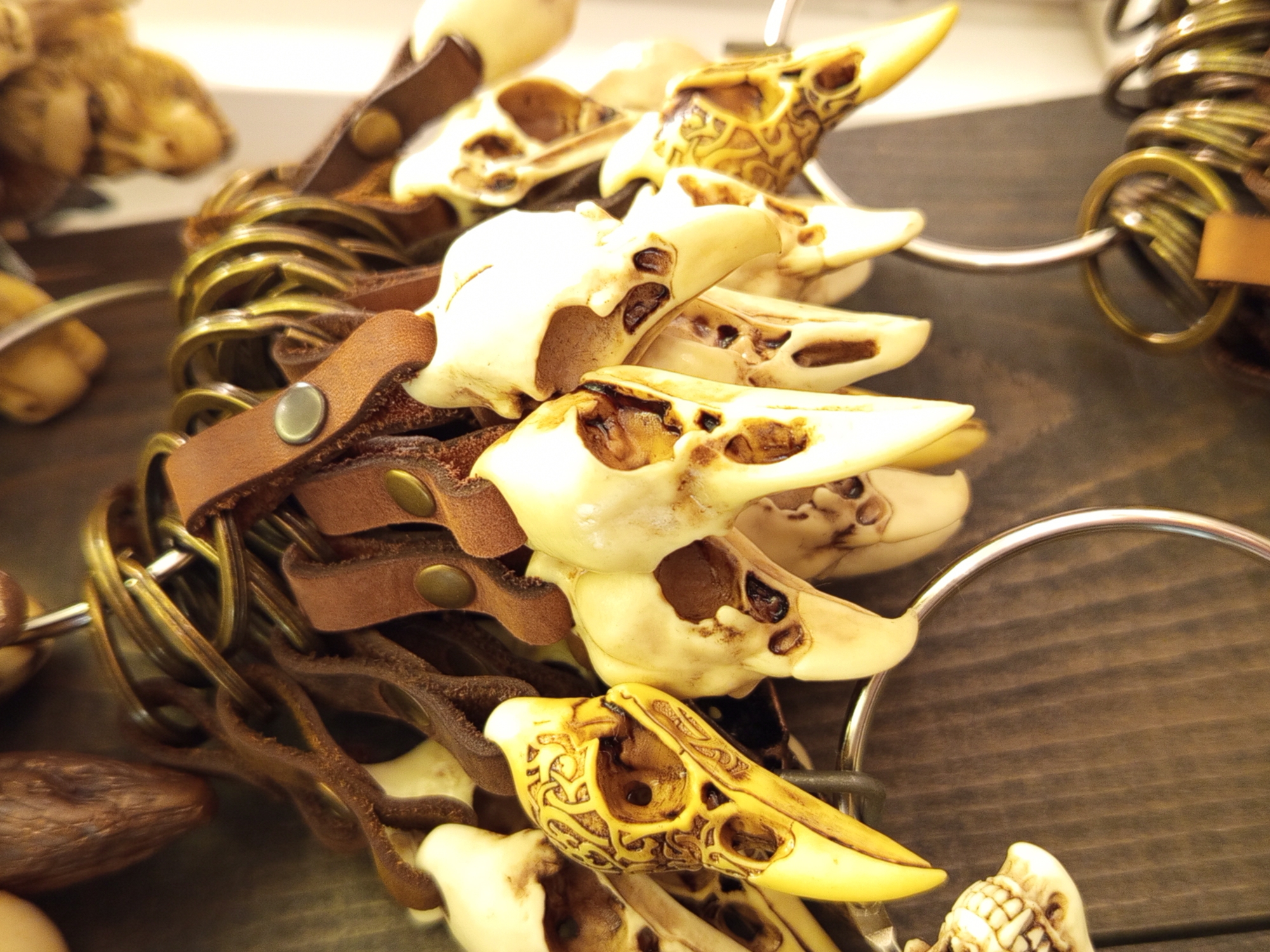 The image shows a close-up, eye-level view of a collection of novelty keychains, each featuring a carved bird skull. The keychains are attached to a large metal ring, which is suspended. The keychains are made of a light-colored material, likely resin or bone, and are detailed with carved patterns and visible eye sockets. Some of the skulls are a pale yellow or cream color, while others have a more golden hue with intricate carvings. They are attached to small, brown leather straps, each secured with a metal snap. These leather straps are also attached to the large metal ring, along with several other smaller metal keyrings. The background is a muted, dark brown wooden surface with vertical grain, suggesting a display shelf or wall in a shop. There are no people or discernible actions taking place. The lighting is somewhat dim, creating soft shadows and highlighting the textures of the carved skulls and leather. The image lacks any specific details that would allow for the identification of a location within Saint Petersburg, Russia.