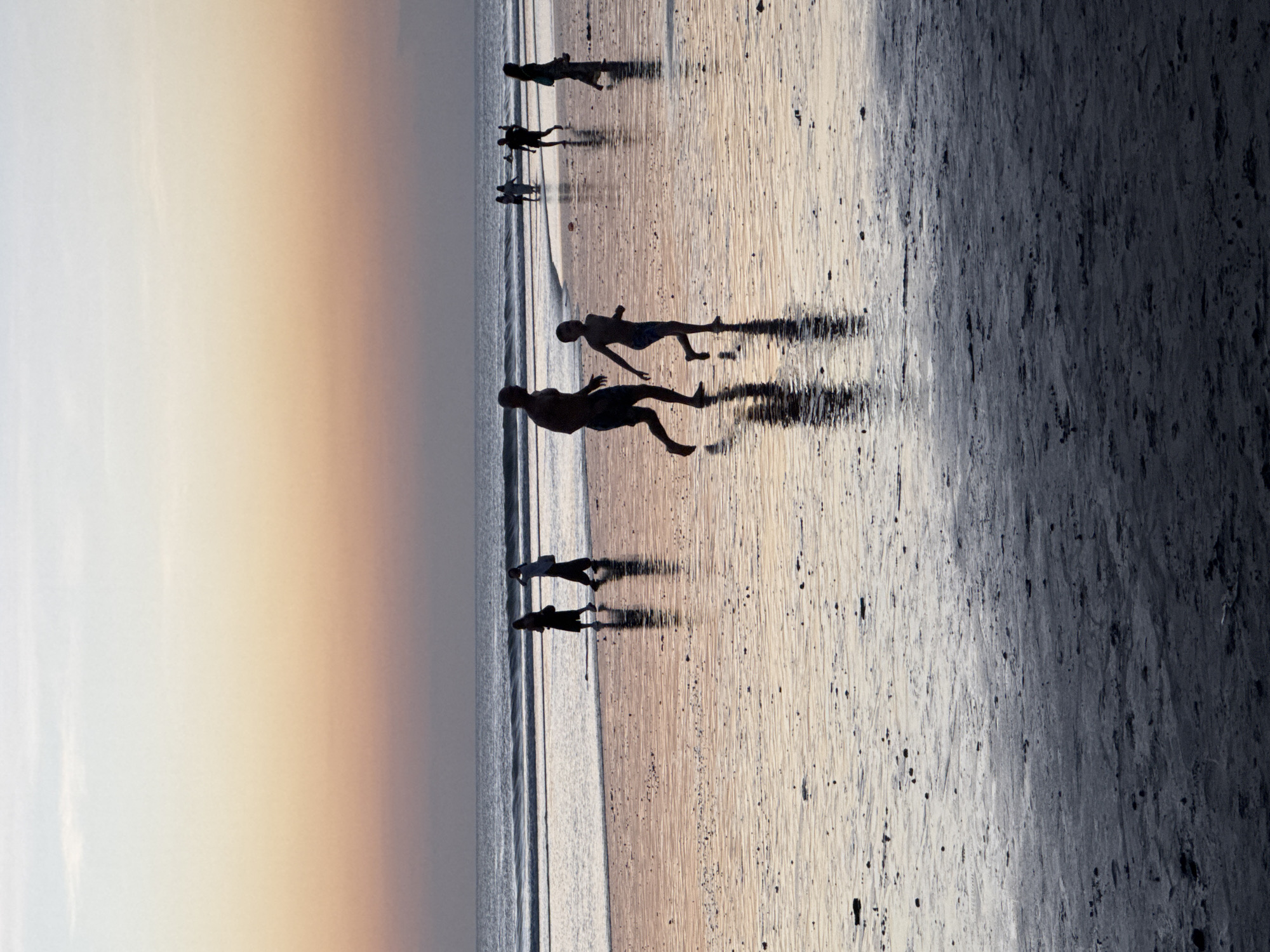 This image depicts a serene sunset scene at Kuta Beach, Bali, Indonesia. Silhouetted figures, including adults and children, are scattered across the wet sand, their reflections shimmering in the shallow water. The sky transitions from a soft orange and pink hue near the horizon to a pale blue overhead, with faint clouds. Gentle waves lap the shore, creating a tranquil atmosphere. The foreground is dominated by the dark, wet sand, dotted with small debris. The overall impression is one of peaceful evening activity on a popular Indonesian beach.