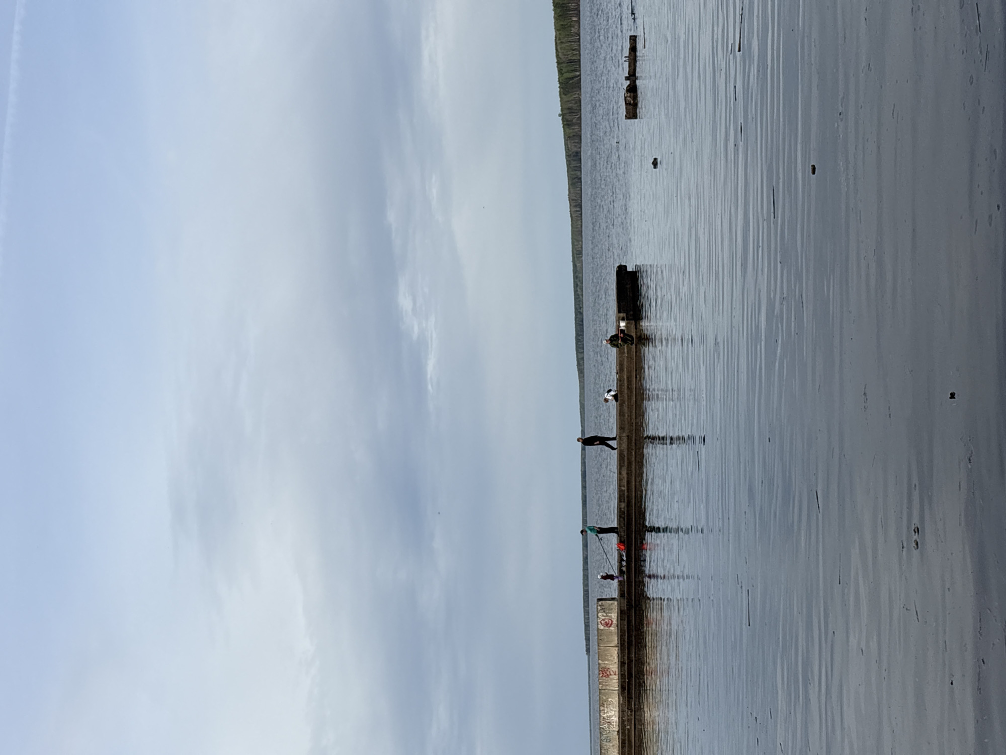 The image depicts a concrete pier extending into a body of water, possibly a lake or river, with a shoreline visible in the background. Several people are present on the pier, some appearing to be fishing. The sky is overcast, suggesting a cloudy day. Debris floats in the water around the pier. Based on the presence of fishing activities and the general appearance of the environment, this location could plausibly be within the city of Chaikovsky, Russia, as it is situated on the Kama River, which provides opportunities for such activities.