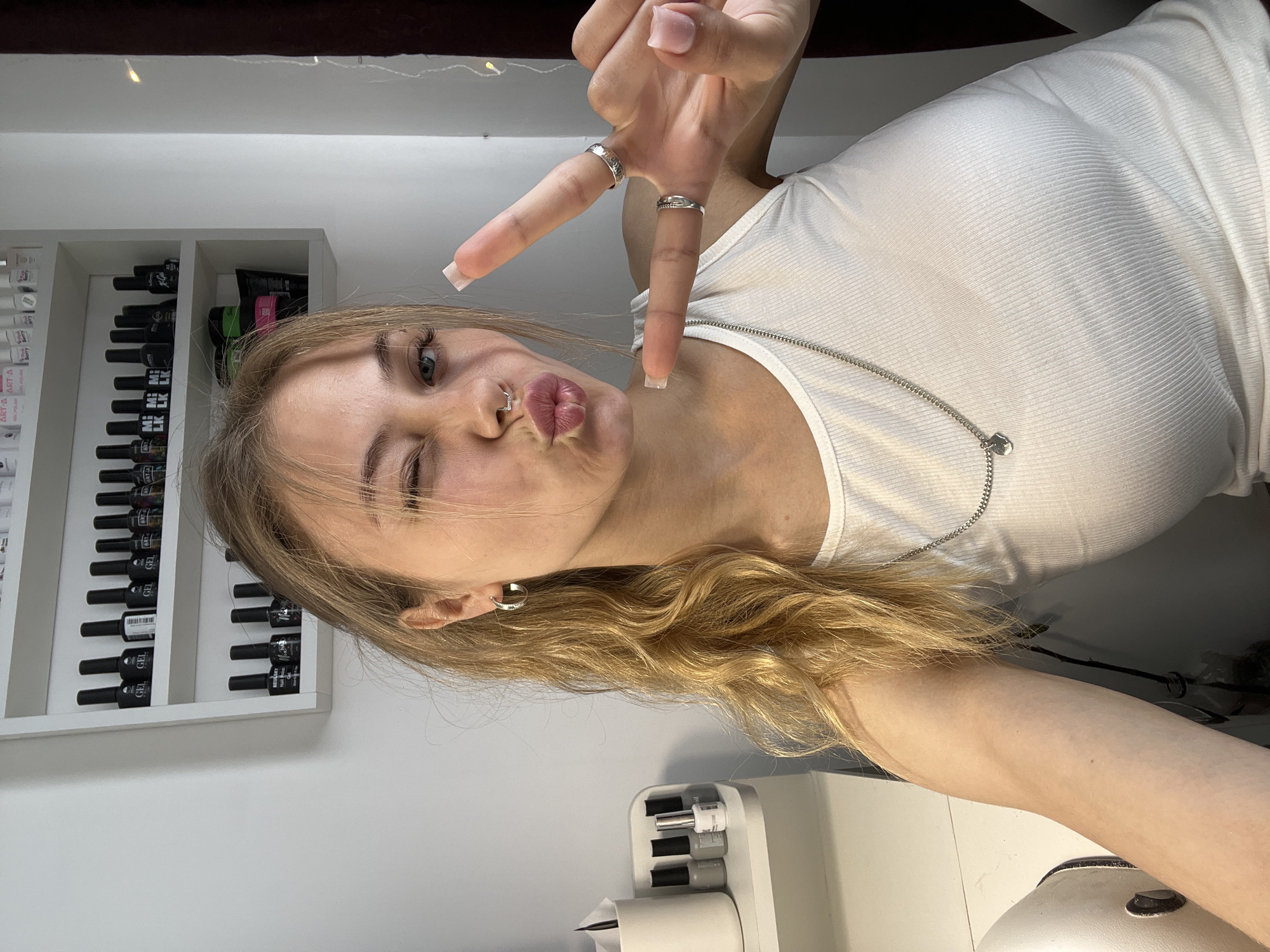 A young woman with blonde hair, wearing a white ribbed tank top and silver jewelry, is posing for a selfie. She has a nose ring and is making a "peace" sign with her fingers. A shelf containing multiple bottles of nail polish is visible behind her. In the foreground is a white manicure table. The location is Фоки, Russia.