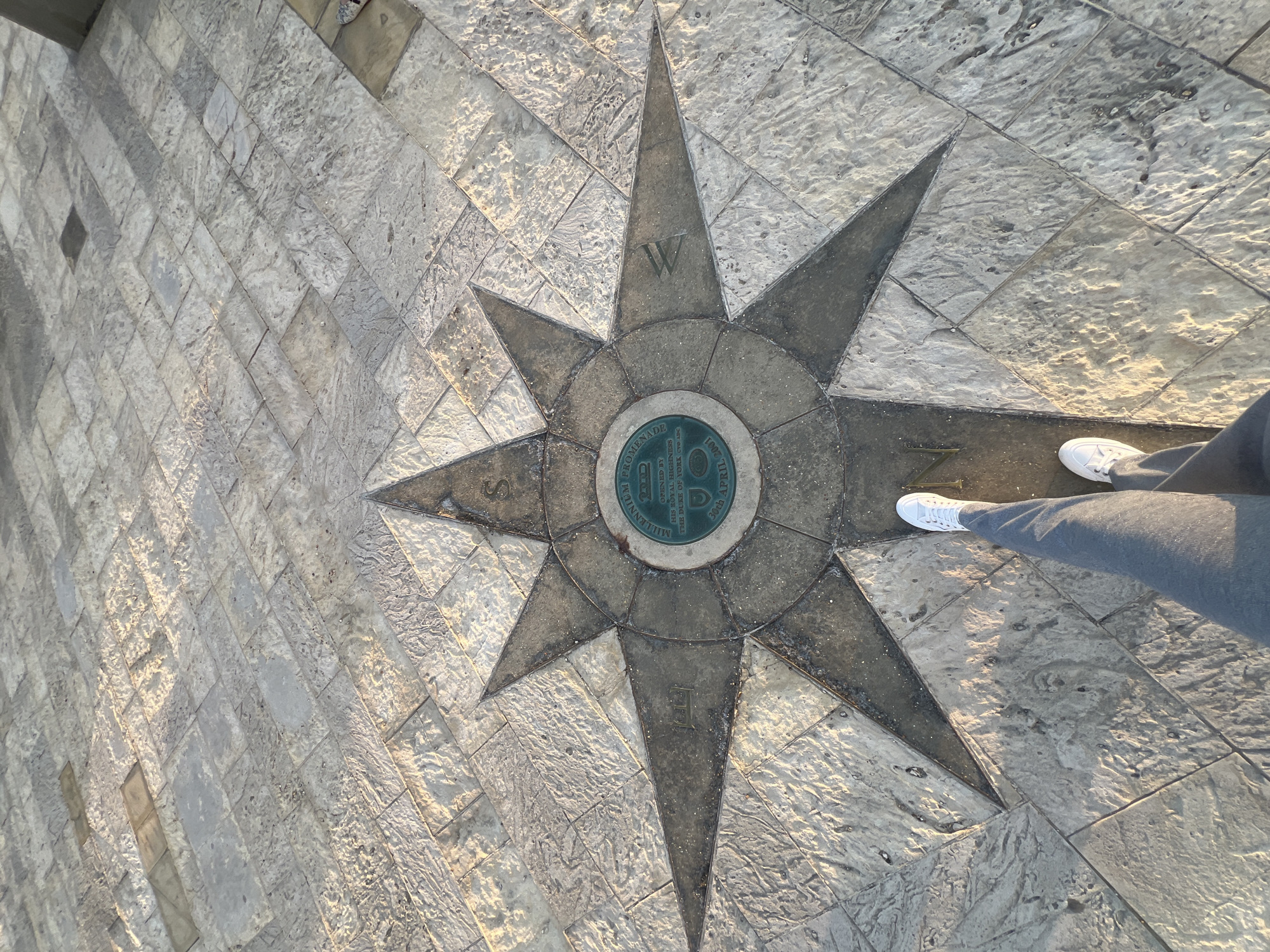 This image depicts a person's legs and feet standing on a paved area featuring a compass rose inlay. The compass rose is made of stone and metal, with an inscription in the center that reads "MILLENNIUM PROMENADE OPENED BY HIS ROYAL HIGHNESS THE DUKE OF YORK 30th APRIL 2001". The paved area is composed of rectangular and square stone tiles arranged in a geometric pattern, reflecting sunlight. The person is wearing grey trousers and white sneakers. The scene is likely located at the Millennium Promenade in Portsmouth, United Kingdom, given the inscription on the compass rose. The lighting suggests it is either early morning or late afternoon.