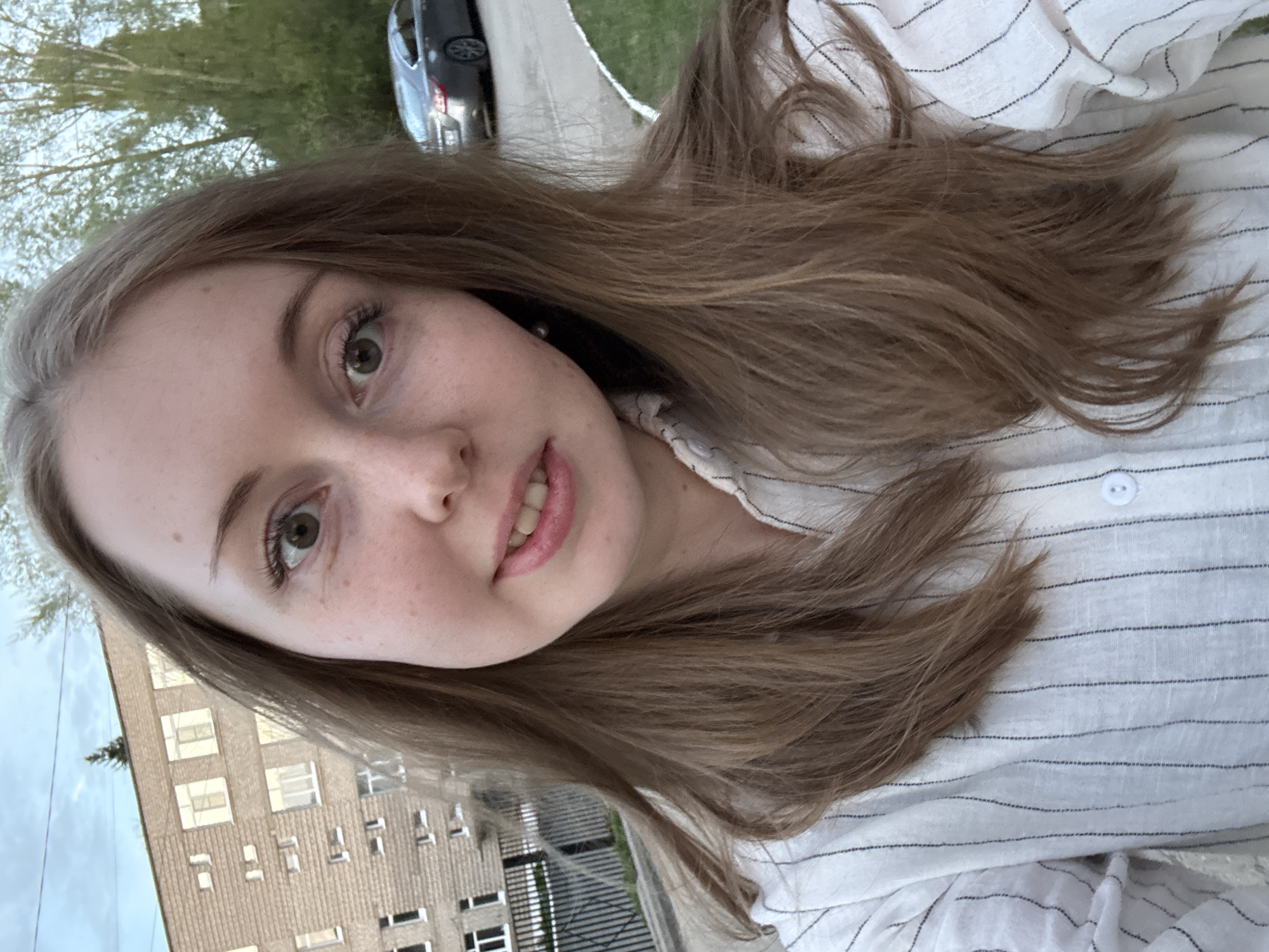 A young woman with light brown hair, fair skin, and freckles is photographed in an outdoor urban setting. She is wearing a white striped shirt. A building with multiple windows and a dark-colored car are visible in the background. The location is the city of Unknown, Unknown.