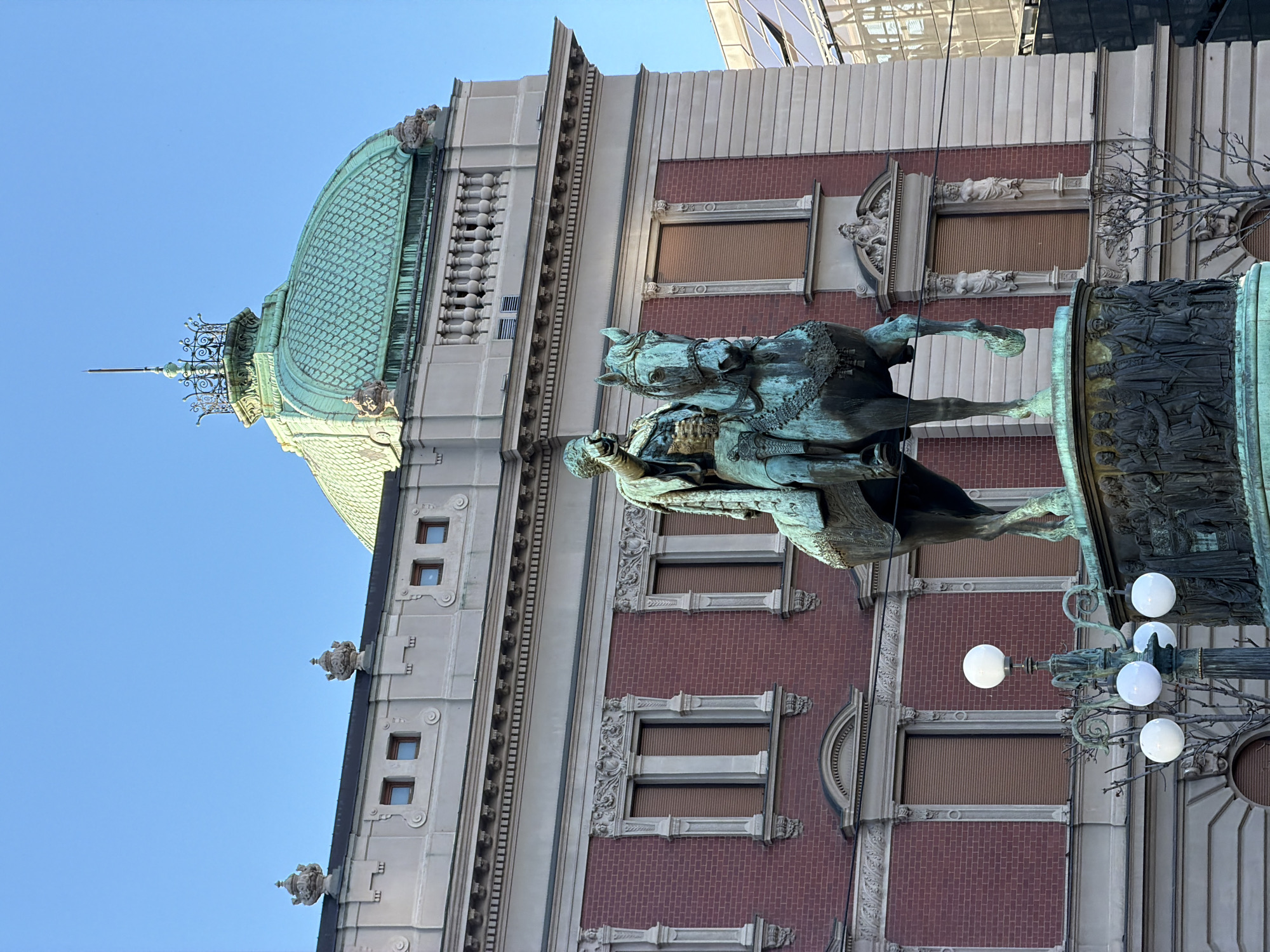 The image shows the monument to Prince Mihailo in Republic Square, Belgrade, Serbia. The equestrian statue of Prince Mihailo is made of bronze. The base of the monument is decorated with reliefs. Behind the monument is the National Theatre with its distinctive copper dome and ornate facade featuring brick and stone detailing. The sky is clear and blue. A modern glass building is partially visible at the top of the frame. An ornate lamppost with several spherical lights is visible in the foreground.