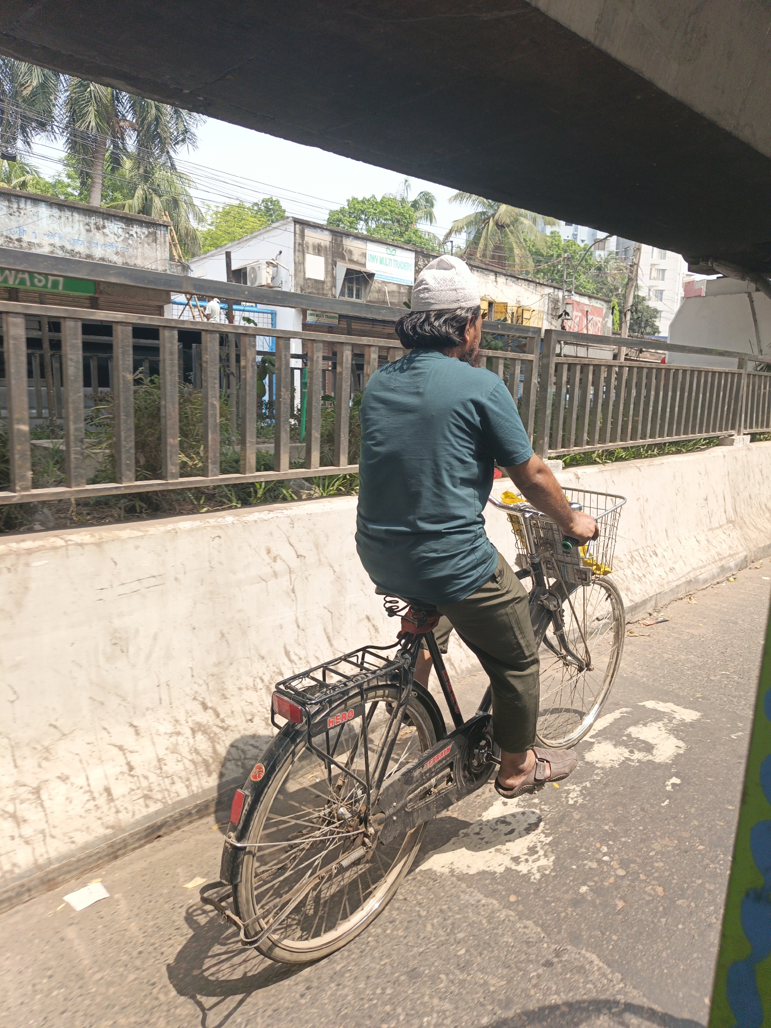 A man rides a black bicycle along a paved road in Dhaka, Bangladesh. He wears a green t-shirt, green pants, brown sandals, and a white skullcap. The bicycle has a basket on the front and a rack on the back, with a Hero brand reflector. The scene is taken from under a bridge. A barrier with railings separates the road from the sidewalk. Buildings and palm trees are visible in the background. "UWU MULTI TRADERS" is visible on one of the buildings.