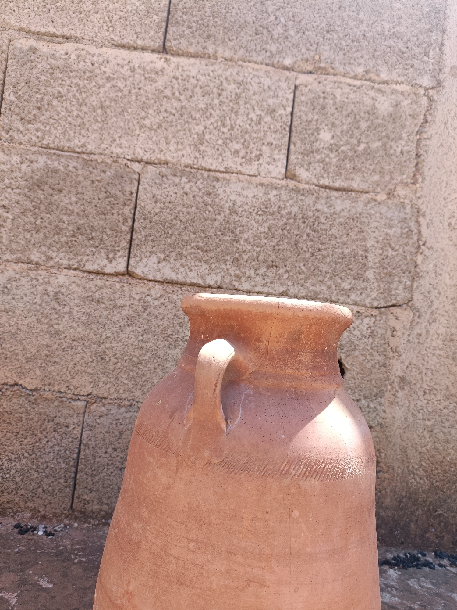 A medium, full-shot captures a large, earthenware pot, placed against a rough, cinder block wall. The pot is a reddish-brown hue and appears old, showing cracks on its upper rim and texture from hand-crafting or age. It features a curved handle attached to its side. The background wall consists of irregularly shaped and aligned cinder blocks, giving a rustic, unfinished aesthetic. The scene is lit with natural sunlight, creating distinct shadows and highlights on both the pot and the wall. The location is Ouaouizert, Morocco. There are no people visible in the scene. The foreground shows part of a concrete or stone ground, with dark pebbles scattered near the base of the wall.
