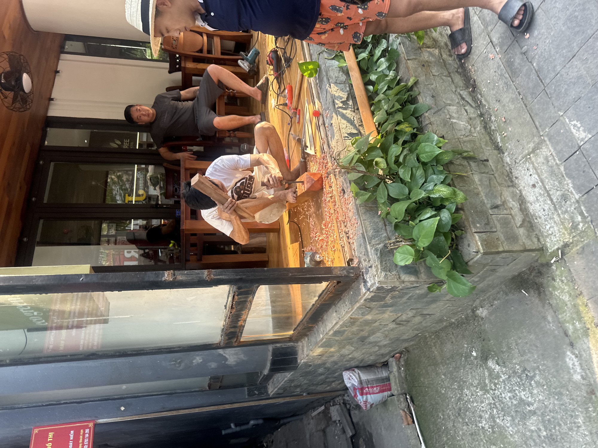 An eye-level outdoor photograph depicts a man carving wood with hand tools on a terrace in Đà Nẵng, Vietnam. The scene is characterized by construction or renovation work.
The man, dressed in a white t-shirt, shorts, and bare feet, is seated, shaping a wooden piece using a chisel and mallet. Wood shavings are scattered around him. Other men are present, one lounging in a chair, another standing partially visible, wearing a hat and patterned shorts. Power tools and additional wood pieces are nearby.
The setting appears to be a shopfront or entrance to a business, evidenced by a window display and a red sign with Vietnamese text. Vegetation grows alongside the work area, adding a natural element to the urban scene. The ground is paved with light-colored bricks and concrete, showing signs of wear. The overall atmosphere suggests ongoing construction or renovation in a commercial area of Đà Nẵng.