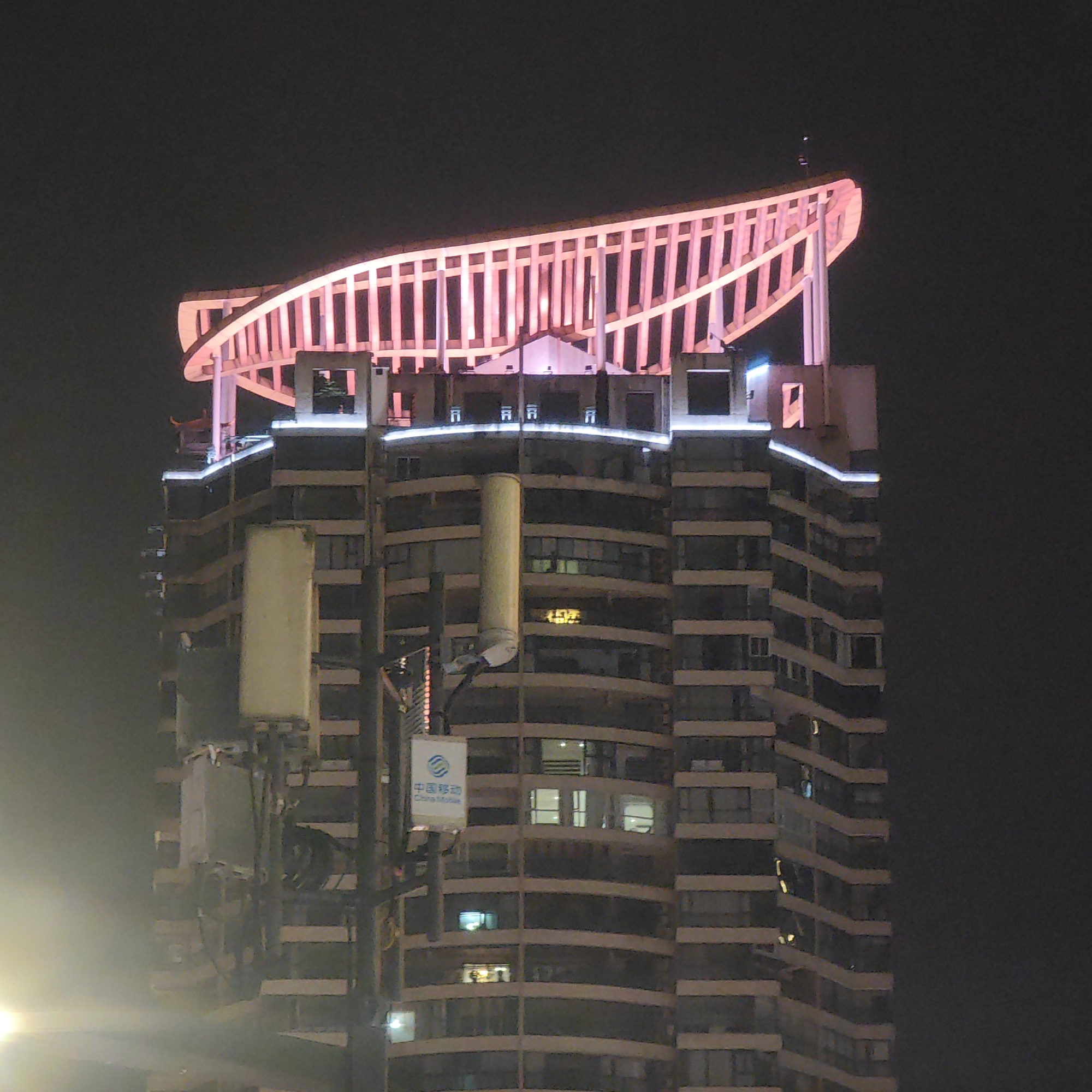 A nighttime shot of a tall building in Yuzhong District, China, possibly a residential or commercial structure, features a distinctive architectural design. The building is lit up with white lights tracing the contours of each floor, and a vibrant pink illuminated structure sits on the roof. Below, a communications tower partially obscures the lower levels, and a sign with Chinese characters and "China Mobile" is affixed to it. The sky is dark, and some fog or haze obscures the base of the building.