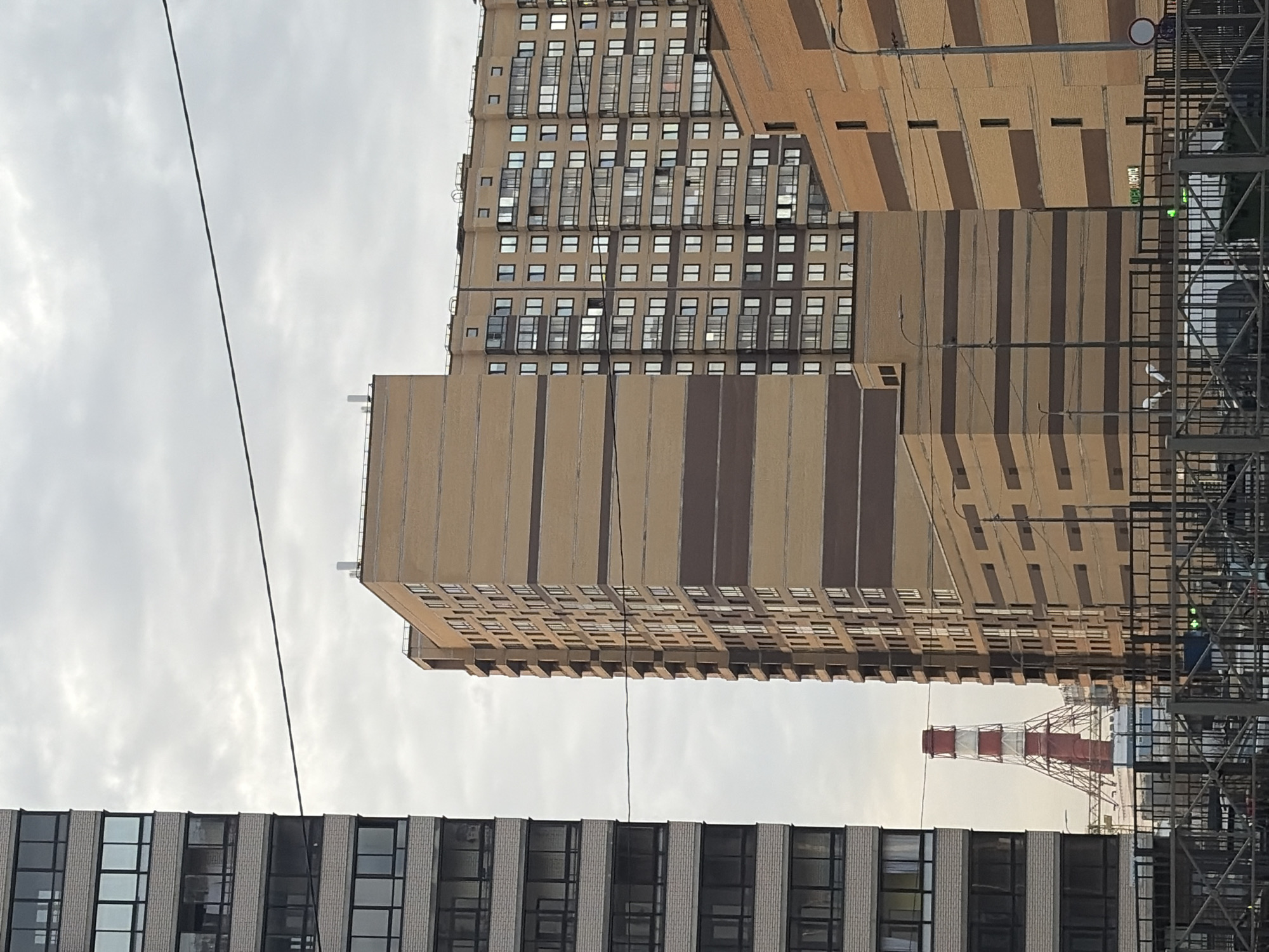The image depicts a cityscape in Saint Petersburg, Russia. Three buildings dominate the frame: two high-rise buildings with a striped brown facade, and a shorter building with a gray-and-white facade. A red-and-white striped smokestack is also visible behind the shorter building. Power lines crisscross the sky in the background, which is filled with a cloudy, overcast sky. A portion of a metal construction scaffolding structure is visible on the right side of the frame. The buildings have numerous windows.