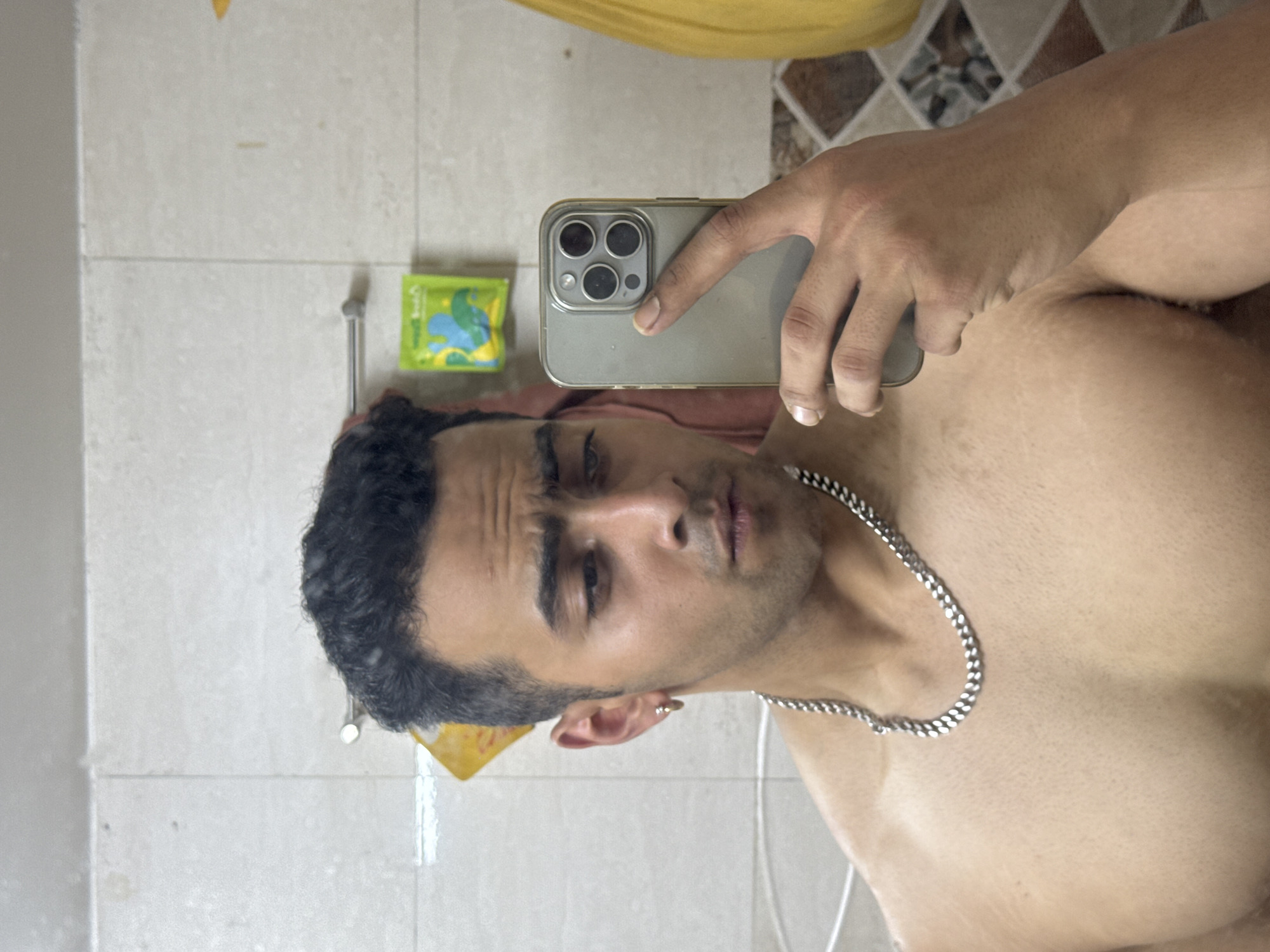 A man takes a selfie indoors, likely in a bathroom given the tiled walls and shower fixtures visible in the reflection. The man has dark curly hair and is wearing a silver chain necklace. He is shirtless. The phone is silver. A package of something is visible on the wall, with blue and green designs. The location is impossible to determine.