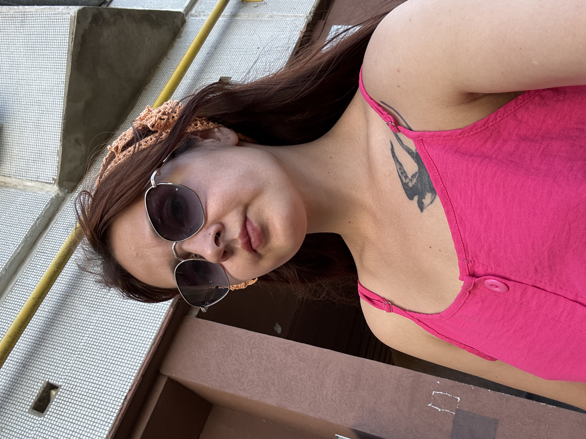 A young woman with long, auburn hair is pictured in a close-up, medium shot outdoors. She is wearing silver-rimmed sunglasses with dark lenses, a light peach crochet headscarf, and a bright pink sleeveless top with a single button closure. A tattoo of a bird in flight is visible on her upper chest. The background shows a textured, light-colored building facade with a yellow pipe running horizontally across it. The building appears to be part of an urban environment. The specific location is identified as Naberezhnye Chelny, Russia, though the precise urban feature is not specified. The lighting suggests it is daytime.