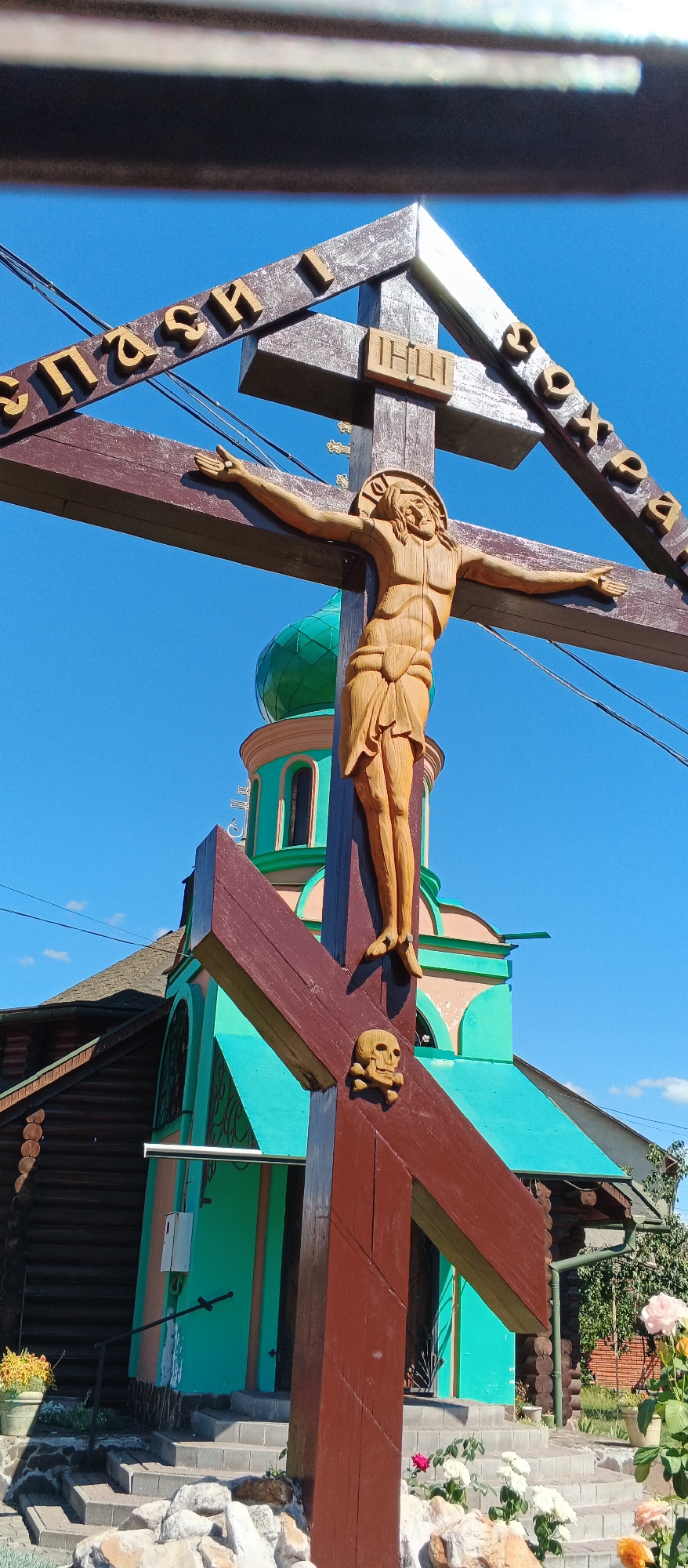 The image shows a wooden crucifix with a carved figure of Jesus Christ. The crucifix is positioned in front of a small building with a green dome and brightly colored walls, which appears to be a church or chapel. The building is made of wood and has a blue-green roof. There is also a second, smaller crucifix with a skull carving at its base. The scene is set outdoors, with a clear blue sky and some sparse clouds visible. In the foreground, there are some rocks and a potted plant. The overall impression is of a religious site or memorial. The image does not depict any people. The inscription on the cross is in Cyrillic script. Based on its architectural style and the presence of a wooden cross, this location is likely a church or a memorial site in a town or village in Ukraine. It is impossible to determine the specific location in Mukachevo, Ukraine, without further context or additional visual cues.