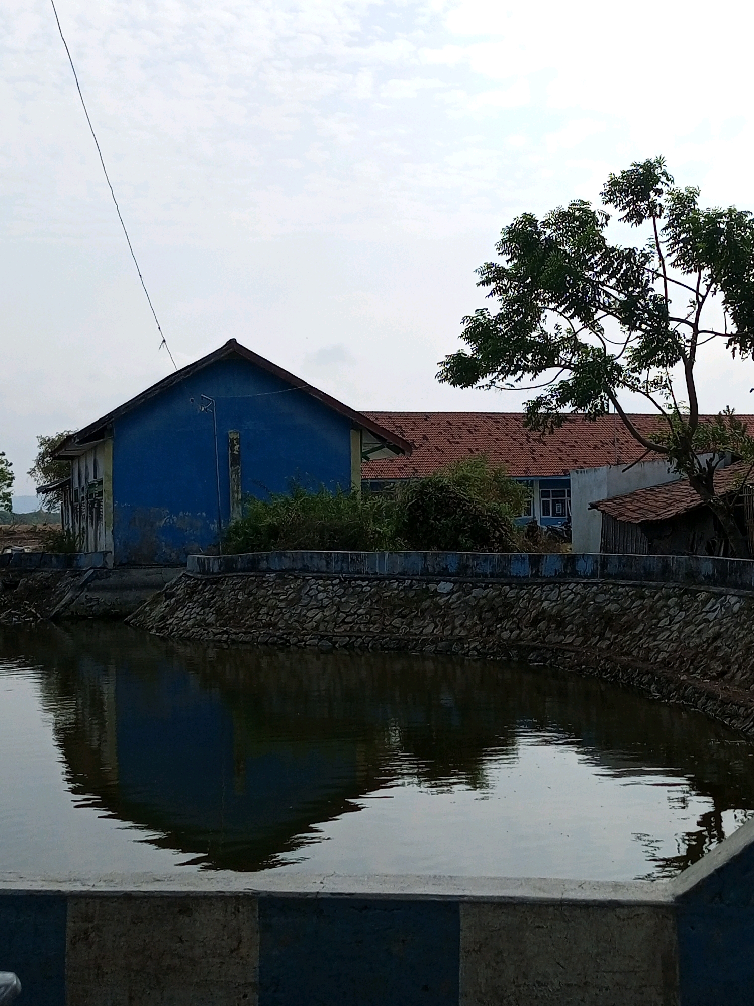 The image depicts a body of water with a reflection of a blue building on the left. The building has a red tiled roof and appears to be weathered. A tree with green leaves is visible on the right, with part of its branches extending over the building's roof. A stone wall runs along the edge of the water, separating it from the building and the surrounding area. The sky is overcast. In the foreground, a concrete structure with blue and gray accents is partially visible. The scene appears to be outdoors, in a rural or suburban setting, possibly a park or a residential area. No people are visible in the image.