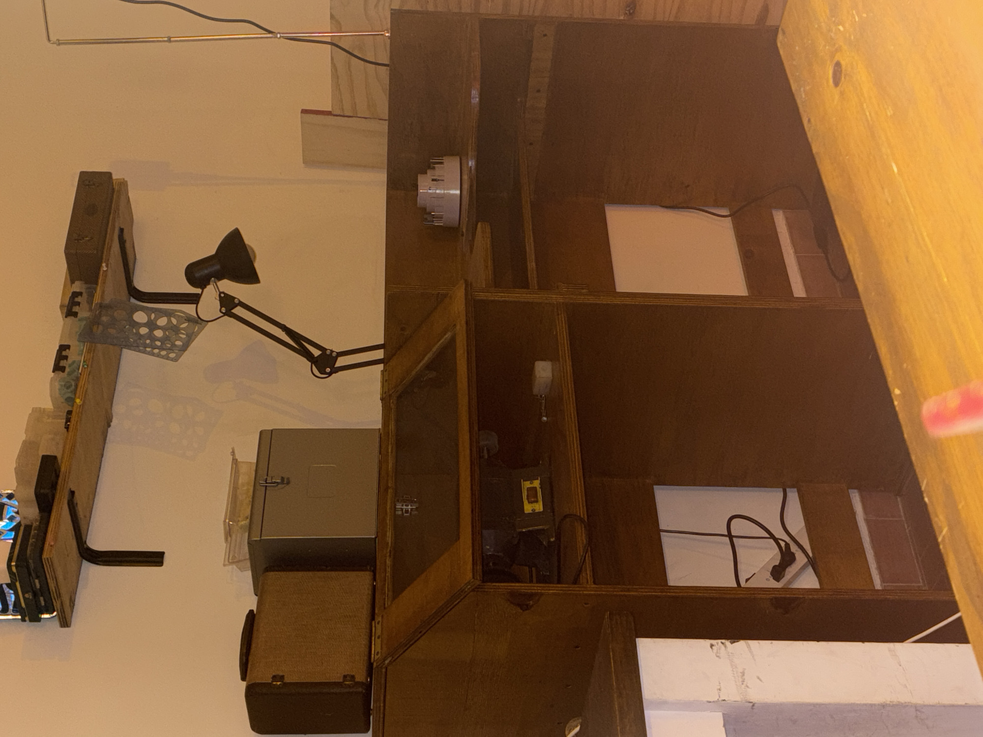 The image depicts a cluttered interior space.
On the left, there is a wall with various objects mounted on it, including what appears to be a shelving unit with electronic components, a desk lamp, and a metal box with a smaller component below.
To the right, there is a large wooden structure, possibly a cabinet or shelving unit, with several open compartments containing electrical wiring and components.
The environment suggests a workshop or storage area. There are no visible people or interactions.
The location in Buenos Aires, Argentina cannot be determined from the image alone.