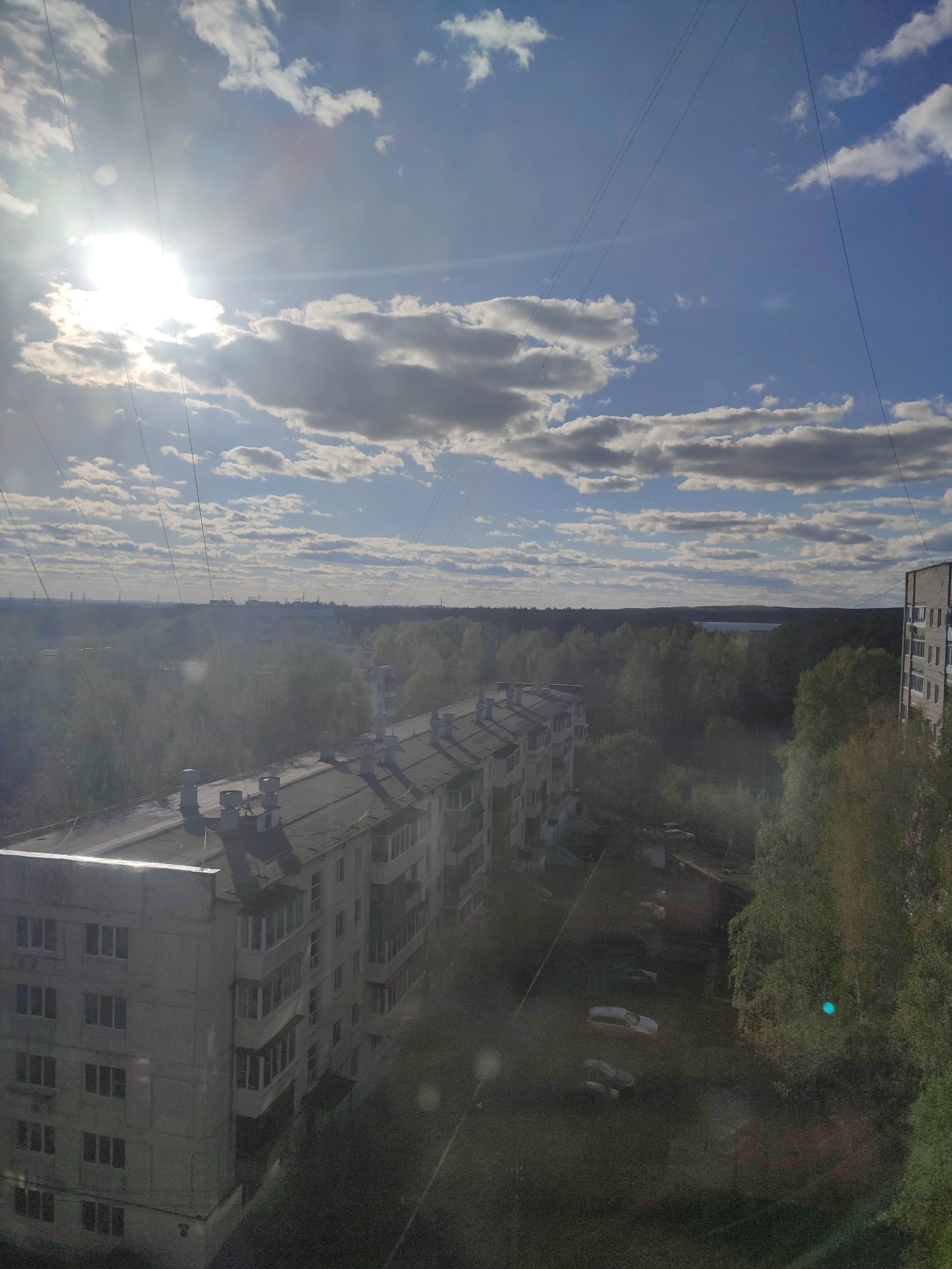 The image shows a view from a high vantage point, possibly a window, overlooking a cityscape of Unknown, Unknown. The scene is dominated by a bright, partially obscured sun in a sky with scattered clouds. Several apartment buildings are visible, characterized by a utilitarian, possibly Soviet-era architectural style, constructed from concrete. The buildings feature flat roofs with visible chimneys or ventilation structures. The surrounding environment is lush with trees, indicating a green, possibly park-like area interspersed within the urban landscape. A thin body of water or lake can be observed in the distance, beyond the treeline. Power lines are strung across the sky, adding to the urban feel. The image is somewhat hazy, and lighting is harsh due to the sun. Several cars can be seen in a parking area on the right-hand side of the shot. The scene appears to be a residential neighborhood with a blend of natural and urban elements.