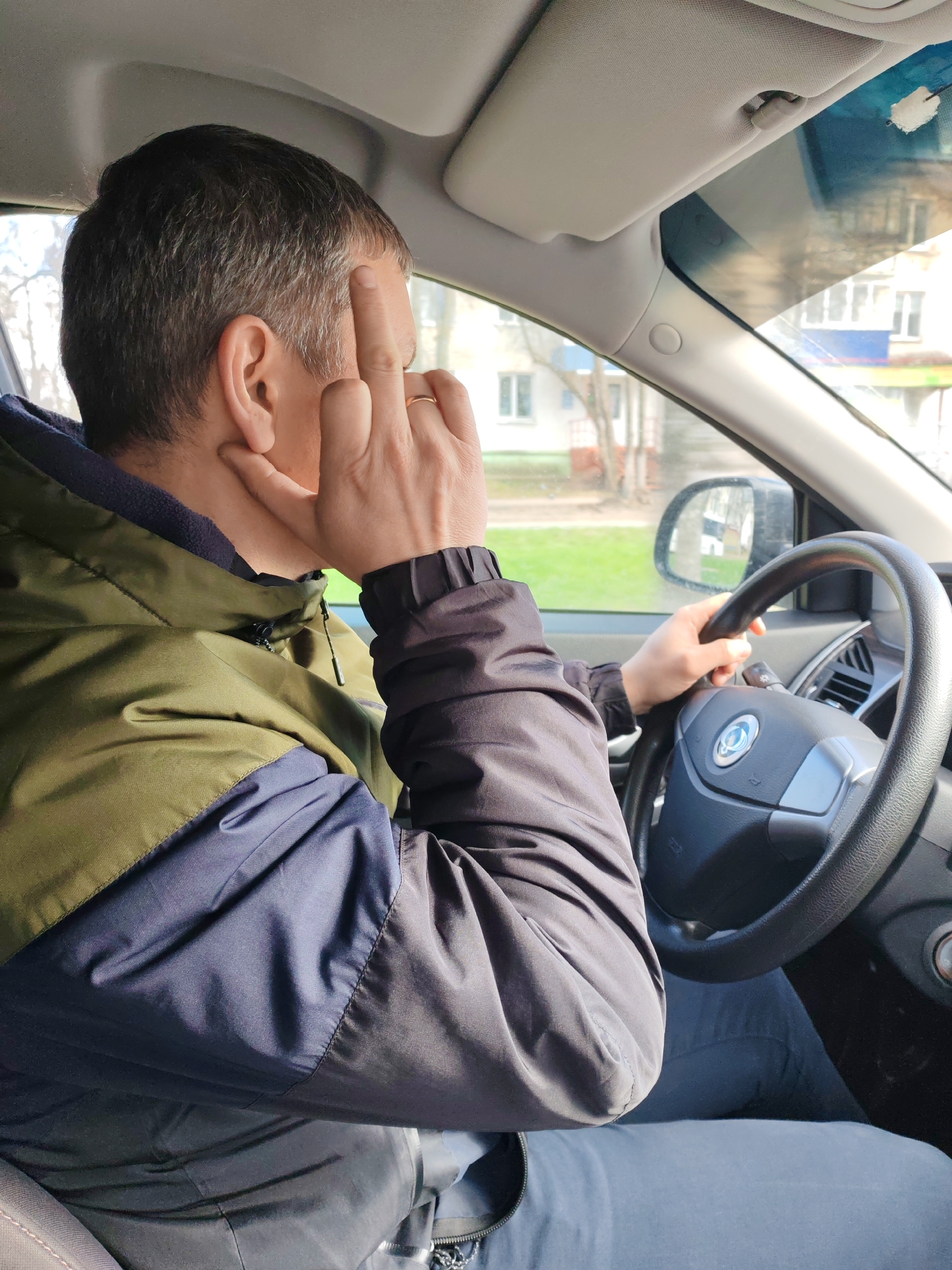 The image shows a man from a rear-right perspective, seated inside a car, likely in the driver's seat. He has short, dark hair with visible grey strands. He is wearing a multi-colored jacket: olive green on the upper back and shoulder, navy blue on the lower back and side, and dark grey/black on the sleeves, which feature elasticated cuffs. A golden ring is present on his right ring finger.
His right hand is raised towards his head, with the middle finger extended and touching his forehead or temple, while the index finger is slightly bent. His left hand is on the car's steering wheel, which has a prominent circular emblem with a stylized design at its center. The car's interior elements, such as the dashboard, sun visor, and part of the A-pillar, are visible.
Through the car's windows, an outdoor scene is visible under bright daylight. The background features light-colored residential buildings with numerous windows, green grass, and some bare trees, suggesting a clear day, possibly in spring or early autumn, in an urban or suburban setting. The location is Chaikovsky, Russia. The man's gesture could imply deep thought, exasperation, or a moment of reflection. There is no visible text in the image.