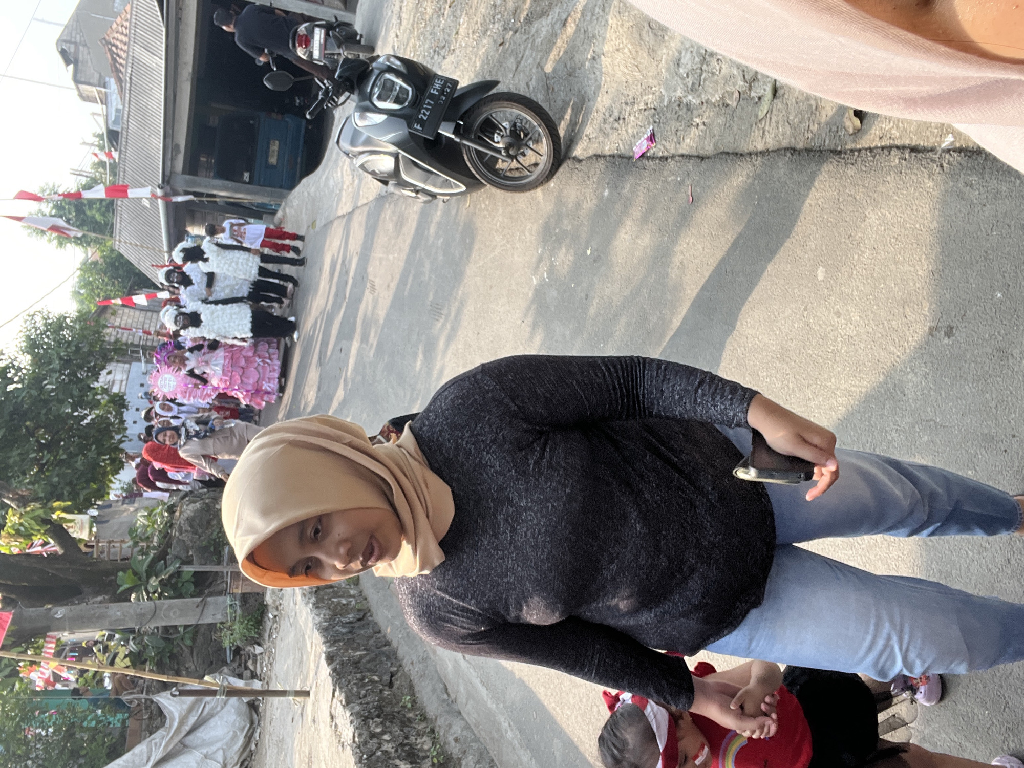 The image depicts a sunny street scene in Klapanunggal, Indonesia, possibly during a local celebration or parade. In the foreground, a woman wearing a beige hijab and a dark grey long-sleeved shirt is walking, holding a cell phone in her right hand. To her left and slightly behind her, a young child in a red top and black skirt, with a white and red headband, is holding her hand.
Further down the street, a group of people are walking, including some individuals in white costumes and others in pink costumes. Red and white Indonesian flags are visible, tied to poles and strung across the street, suggesting a national holiday or event. A dark-colored scooter is parked on the right side of the road. Buildings with corrugated metal roofs line the street. The ground is paved with asphalt and concrete. The overall atmosphere appears to be festive and community-oriented.