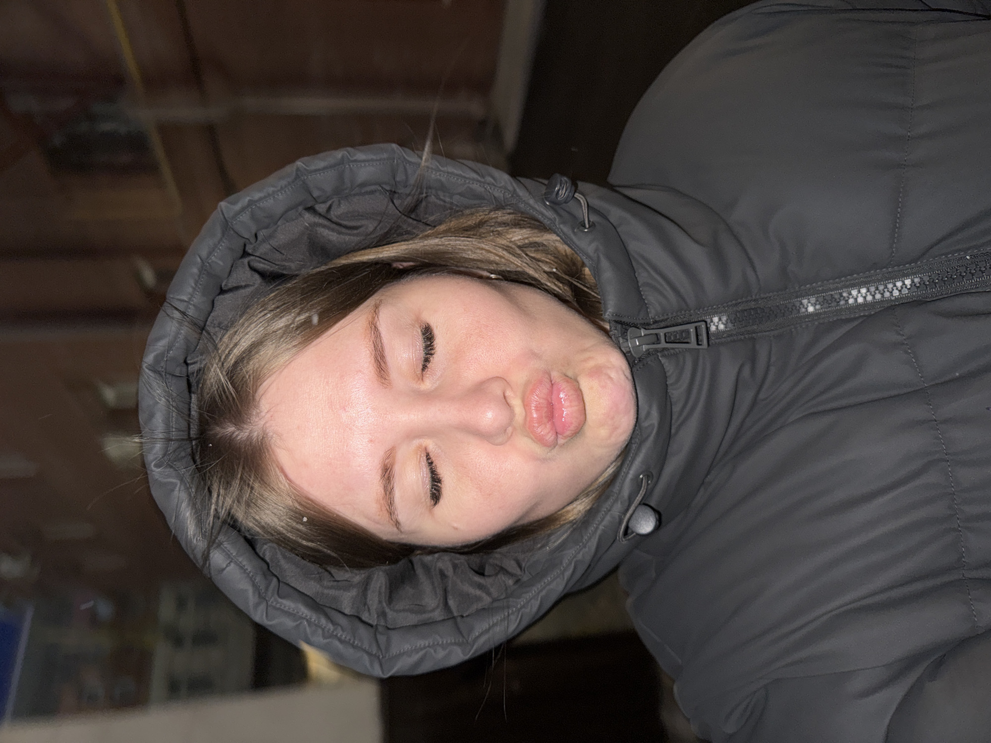 The image presents a close-up portrait of a young female, oriented at a tilt. She is wearing a dark grey or black puffer jacket with the hood pulled up around her face. Her eyes are closed, and her lips are pursed into a playful "fish face" expression. Light brown hair frames her face, with a few strands visible.
Small white specks, consistent with snowflakes, are visible on her hair, eyebrows, and the outer fabric of her jacket, indicating current snowfall or recent exposure to snow. The background is dark and heavily blurred, showing indistinct shapes and faint linear structures, suggesting a nighttime outdoor setting or a dimly lit, possibly industrial, indoor environment. The overall lighting is dim. A zipper and a drawstring toggle are visible on her jacket. The subject's expression conveys a sense of lightheartedness or playfulness. No visible text is present in the image. The context for the location is Perm, Russia.