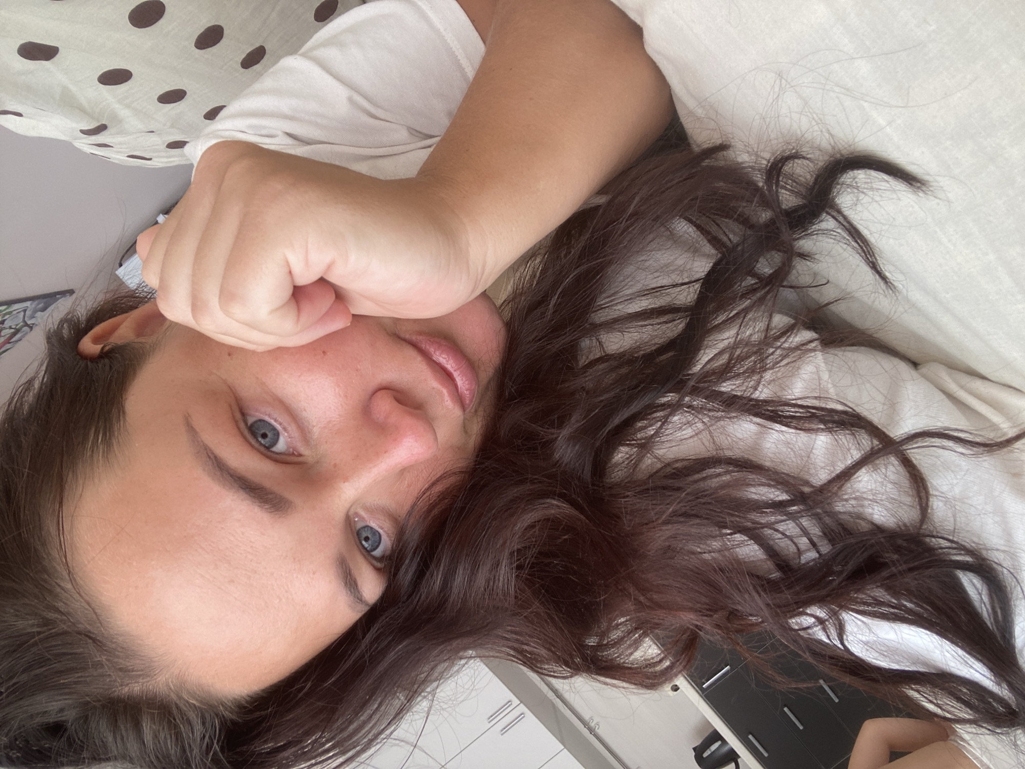 A woman with long, dark brown hair and blue eyes rests her head on her hand. She is wearing a white t-shirt and is lying on a bed with a white duvet and a pillow with brown polka dots. The background shows part of a kitchen with white cabinets and dark drawers. The scene appears to be indoors.