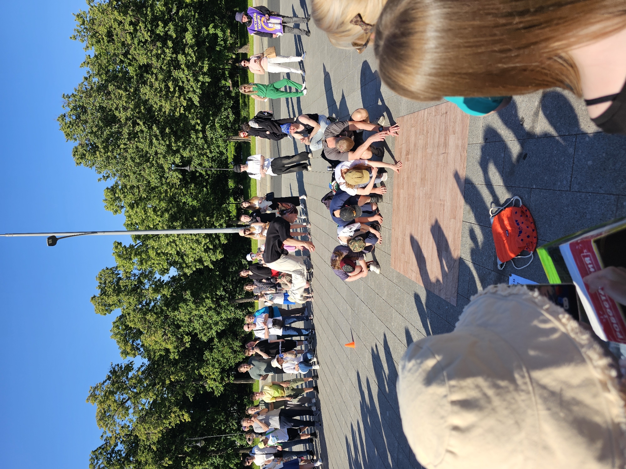 This image captures an outdoor street performance or practice session in an urban public space in Moscow, Russia, characterized by the Russian text "ВЫСТАВКА КОШЕК" (CAT EXHIBITION) visible on a participant's vest. The scene is illuminated by bright sunlight under a clear blue sky, casting distinct shadows on the light grey paved ground.
In the immediate foreground, the top of a light-colored bucket hat is visible on the left, belonging to a person holding a book. To the right, the back of a person's head and shoulders with long brown hair, wearing a black top, is visible, also facing away from the camera.
The central focus is a group of approximately six individuals performing on two light brown, wood-patterned mats laid on the pavement. Most are in a crouched or bent-over position, hands on the ground, forming a line. One performer in a black shirt is captured mid-air, upside down, executing an acrobatic flip or handstand. Another individual, wearing a black tank top and blue bandana, sits nearby on the pavement, observing the active performers.
Behind the performers, a crowd of approximately 20-30 spectators stands in a semi-circle. They are casually dressed, and many are holding smartphones to record or photograph the event. On the far right of the spectator group, a person wears a purple vest with "ВЫСТАВКА КОШЕК" printed in white Cyrillic text, holding two white rectangular signs or objects.
The background consists of dense foliage from numerous mature green trees, suggesting a park or tree-lined avenue. Two grey lampposts, one with a modern LED fixture, punctuate the upper frame. A small orange traffic cone is visible on the pavement to the left of the performers. A red drawstring bag rests on the pavement in the mid-foreground.