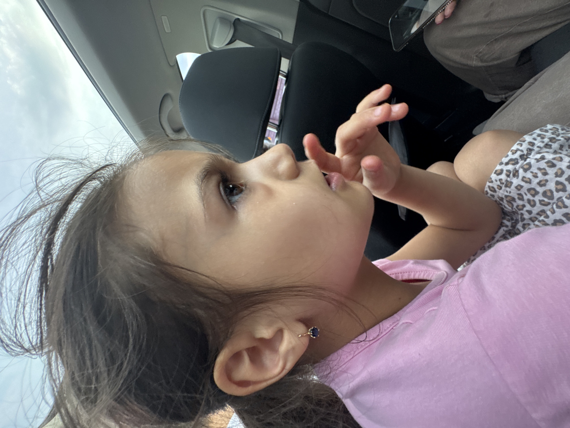 A young girl with dark brown hair, styled in a ponytail with visible flyaways, is seated in the passenger compartment of a vehicle in Suprunivka, Ukraine. She is depicted in profile, facing right, with her right hand raised, index finger touching her lips, suggesting contemplation. Her visible left ear features a gold-colored earring with a dark blue or purple stone. She wears a light pink short-sleeved top with a collar and a white garment with a brown leopard print pattern. The vehicle's interior includes a black headrest, a light gray roof lining, and a sunroof, through which an overcast sky is visible. In the lower right corner, the legs of another individual, clad in light brown trousers, are partially visible, alongside a hand holding a black smartphone.