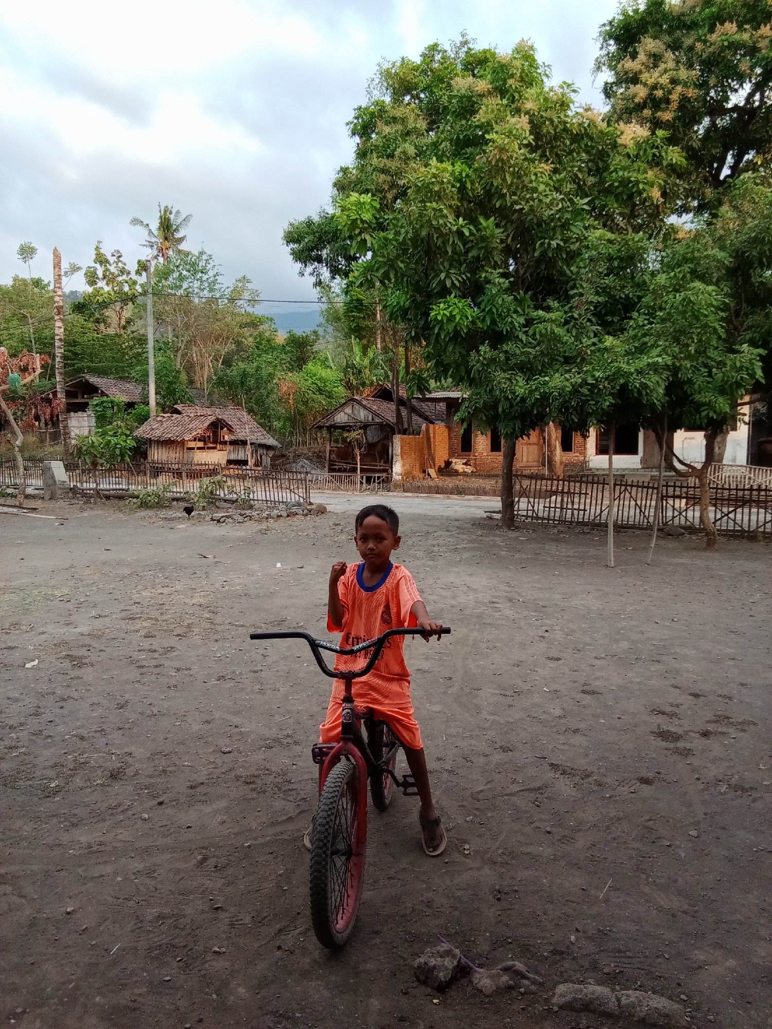 A young boy, wearing an orange t-shirt and shorts, is seated on a red and black bicycle in what appears to be the village of Selowogo, Indonesia. He is facing the camera with his left fist raised. The ground is dirt and gravel. Behind him, there are several small wooden structures with thatched roofs, surrounded by a low wooden fence. Large green trees with some flowering branches are visible in the background, along with a cloudy sky. The overall scene suggests a rural, undeveloped environment.