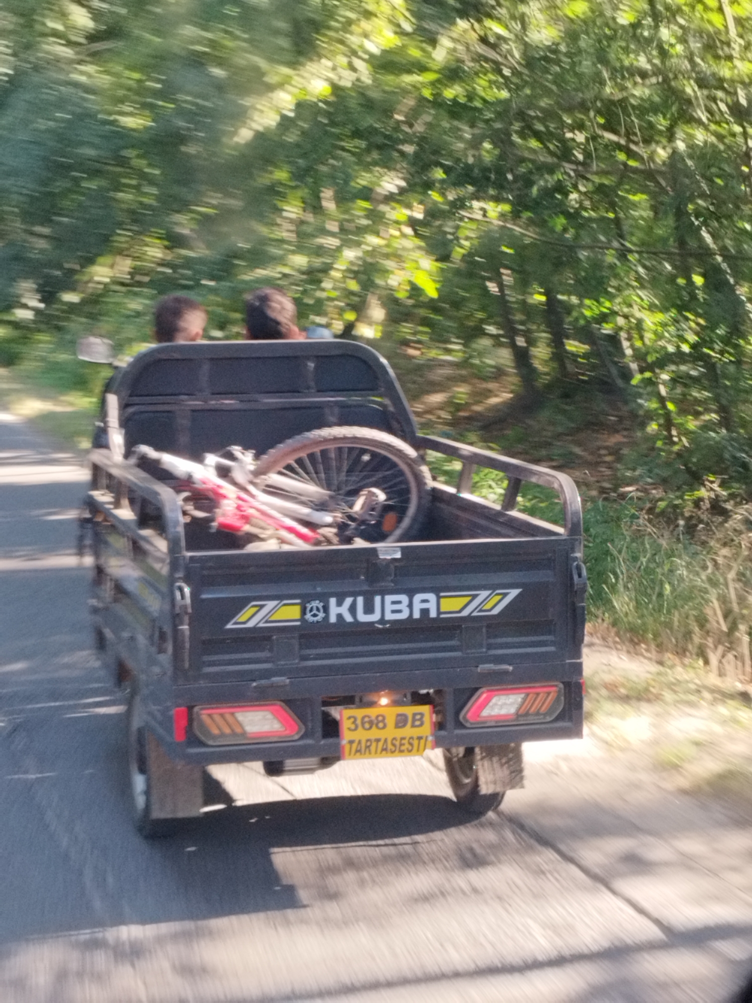 A three-wheeled vehicle, likely a three-wheeled utility vehicle, is traveling on a paved road in Săbăreni, Romania. The vehicle has a black bed that contains a disassembled bicycle with a red frame and silver wheels, and two individuals are seated in the vehicle's cabin. The vehicle is branded with "KUBA" logo. The surrounding environment features lush green trees and vegetation, suggesting a rural or semi-rural setting. The road surface appears worn, and shadows are cast on it, indicating a sunny day. The image is taken from a moving perspective, causing some motion blur, particularly in the background vegetation and the road. A license plate with the numbers "368 DB" and text "TARTASESTI" is visible on the rear of the vehicle.