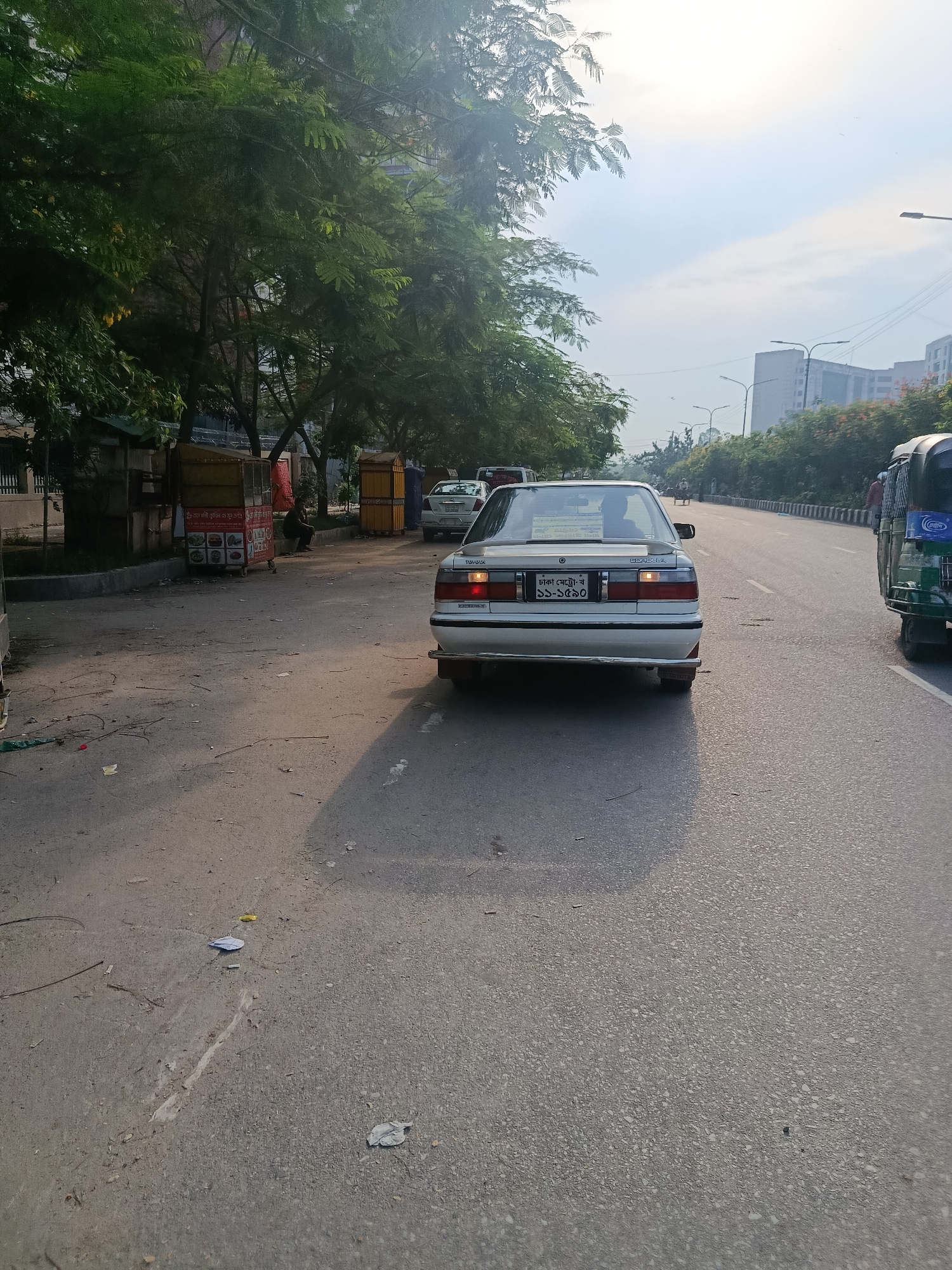 The image shows a street scene in Dhaka, Bangladesh. A white Toyota Corolla, license plate ঢাকা মেট্রো-ঘ ১১-১৫৯০, is in the foreground, facing away from the viewer on a two-lane road. To the right of the Corolla is a CNG auto rickshaw. On the left side of the road, there is a sidewalk with trees, stalls, and people. The road appears to be asphalt with some litter. Tall buildings can be seen in the background under a partly cloudy sky.