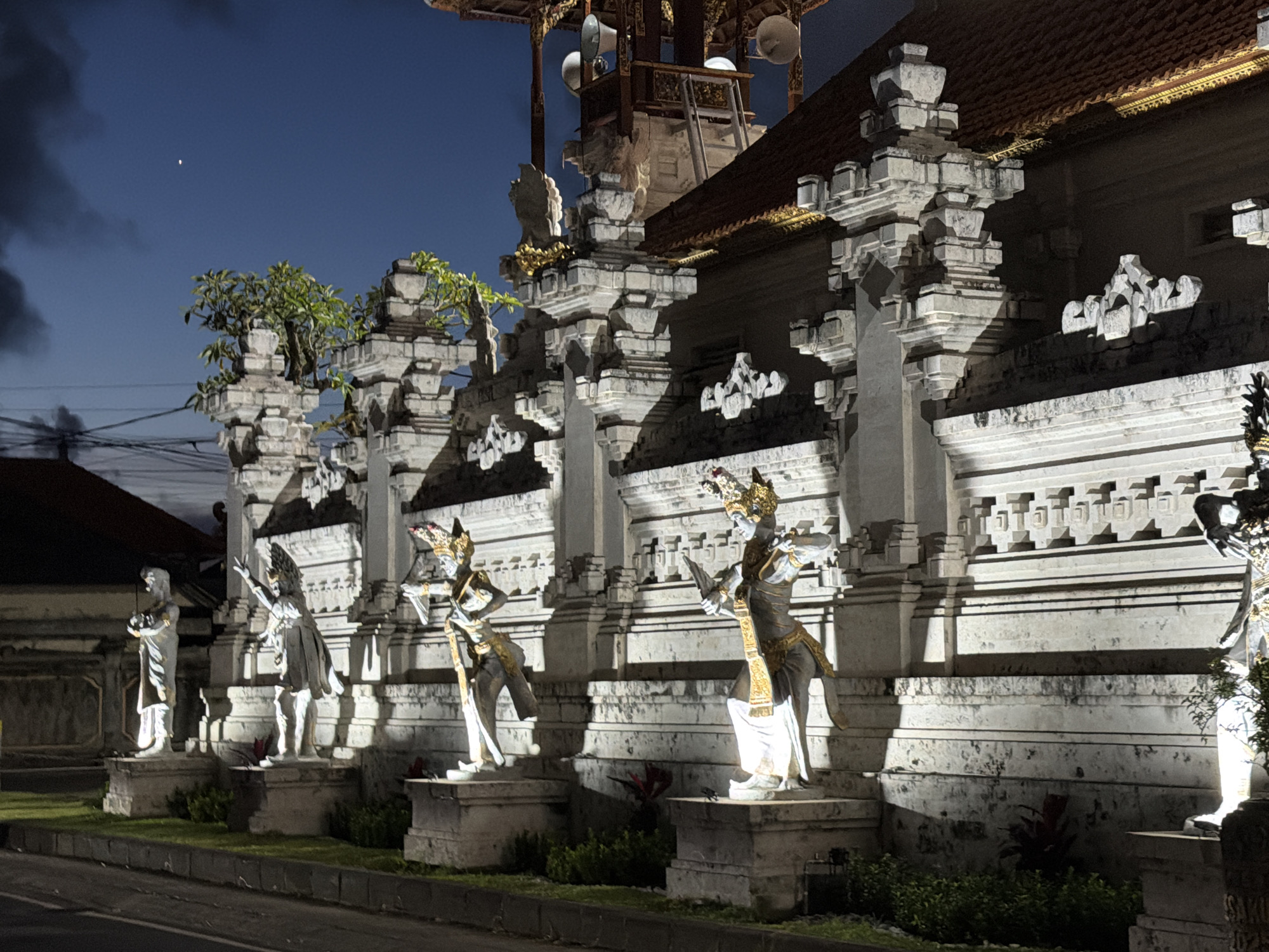 The image shows an outdoor scene in Kutuh, Indonesia, at night. The prominent features are a row of five statues in traditional Balinese style, illuminated by a light source not visible in the image. The statues are positioned along the front of a building with ornate stone architecture, featuring repetitive decorative elements and tiers. The building has a tiled roof. Behind the building, a structure with a platform is visible, possibly for loudspeakers. In the foreground, there's a narrow strip of grass and a curb bordering a road. The sky is a gradient of blue to dark gray, suggesting either twilight or a partially overcast night. The lighting is predominantly artificial, highlighting the statues and architectural details. A building is faintly visible in the background left, obscured by the darkness. There are no people directly visible other than the statues themselves.