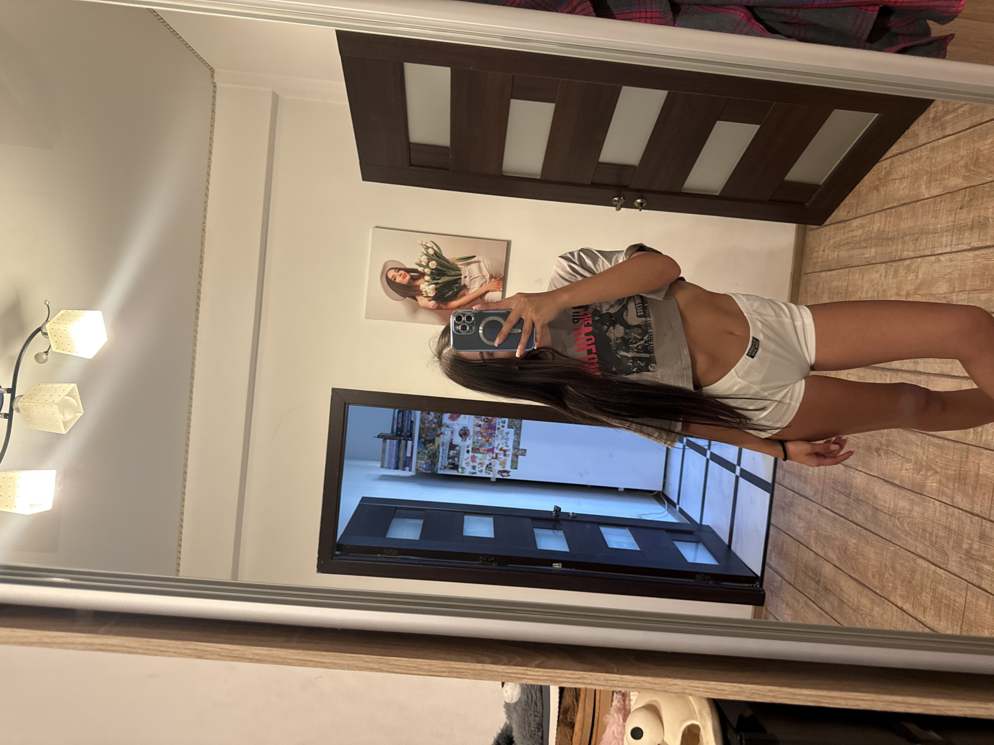 The image shows a young woman taking a mirror selfie in what appears to be a residential interior, possibly an apartment or house. The woman has long dark hair and is wearing a grey t-shirt and white shorts. She is holding a smartphone to take the picture. In the background, there is a wooden floor, a dark brown door with glass panels, and a painting on the wall. There is also a large mirror reflecting the scene. The lighting suggests it is daytime, with light coming from overhead fixtures. There are no clear indications of the specific location within Chișinău, Moldova, or any city.