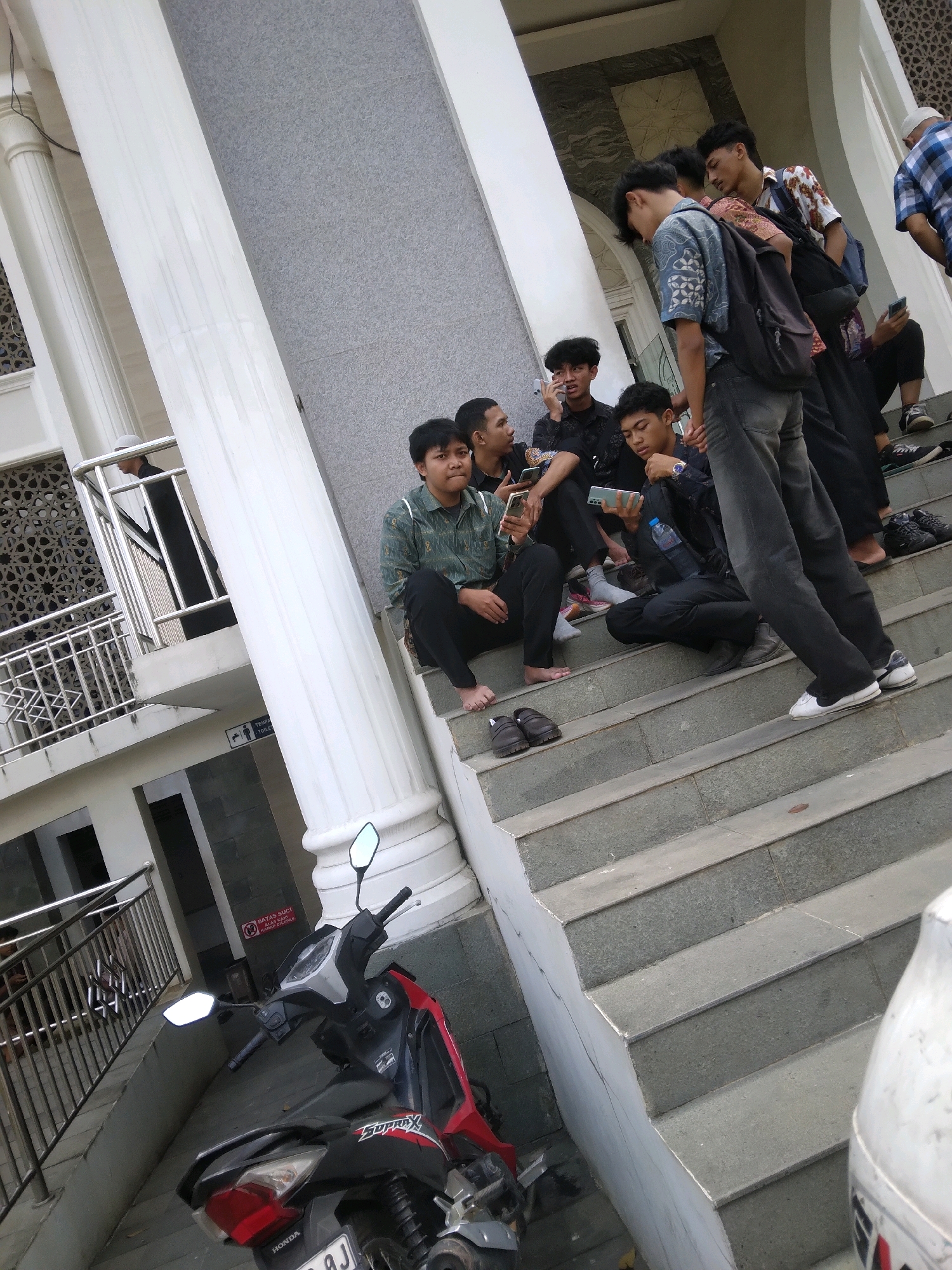 A group of young men, appearing to be teenagers or young adults, are gathered on the steps of a building, possibly a mosque or community center, in Sokaraja, Indonesia. The architectural style suggests a tropical climate with large, white pillars and decorative screen work.
Several individuals are seated and standing on the gray concrete steps. Two men are looking at their phones, one holding a smartphone and another holding a longer device. Another man is wearing a backpack and looking down. Others are casually interacting, with one man gesturing with his hand. The men are dressed in a variety of casual attire, including shirts, t-shirts, and pants. Some are barefoot, while others wear shoes.
In the foreground, a red and black motorcycle is parked at the bottom of the steps. The motorcycle has mirrors and appears to be a common model in the region. The background shows more of the building's structure, including a railing and more architectural details. A sign with Indonesian text is visible on the wall. The overall scene depicts a moment of relaxation and social interaction among young men in an outdoor urban setting.
