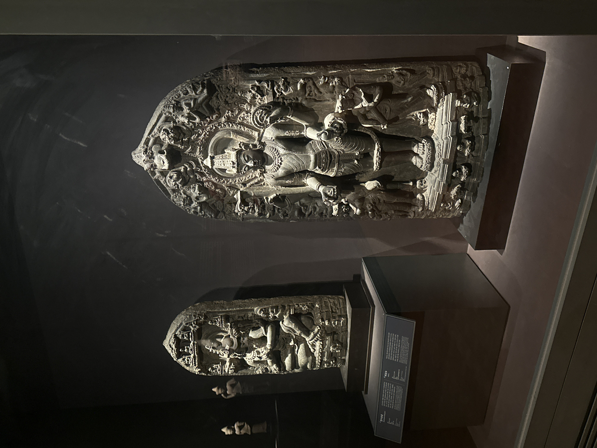 The image shows two ancient stone sculptures displayed in a museum setting. The sculpture on the right is larger and more intricate, depicting a multi-armed deity with a headdress, elaborate jewelry, and detailed clothing. Surrounding the central figure are smaller figures and decorative elements. The sculpture on the left is smaller and appears to be a seated figure, also with multiple arms and adorned with headdress and jewelry. Both sculptures are placed on dark pedestals, and the background is dark, suggesting a museum exhibit designed to highlight the artifacts. In the background, on the left, a third, smaller sculpture is partially visible. A descriptive plaque is placed in front of the left sculpture. The lighting in the scene is focused on the sculptures, with the surrounding area in shadow. The overall presentation suggests a cultural heritage exhibition. This image was taken at the National Museum of Korea in Seoul, South Korea.