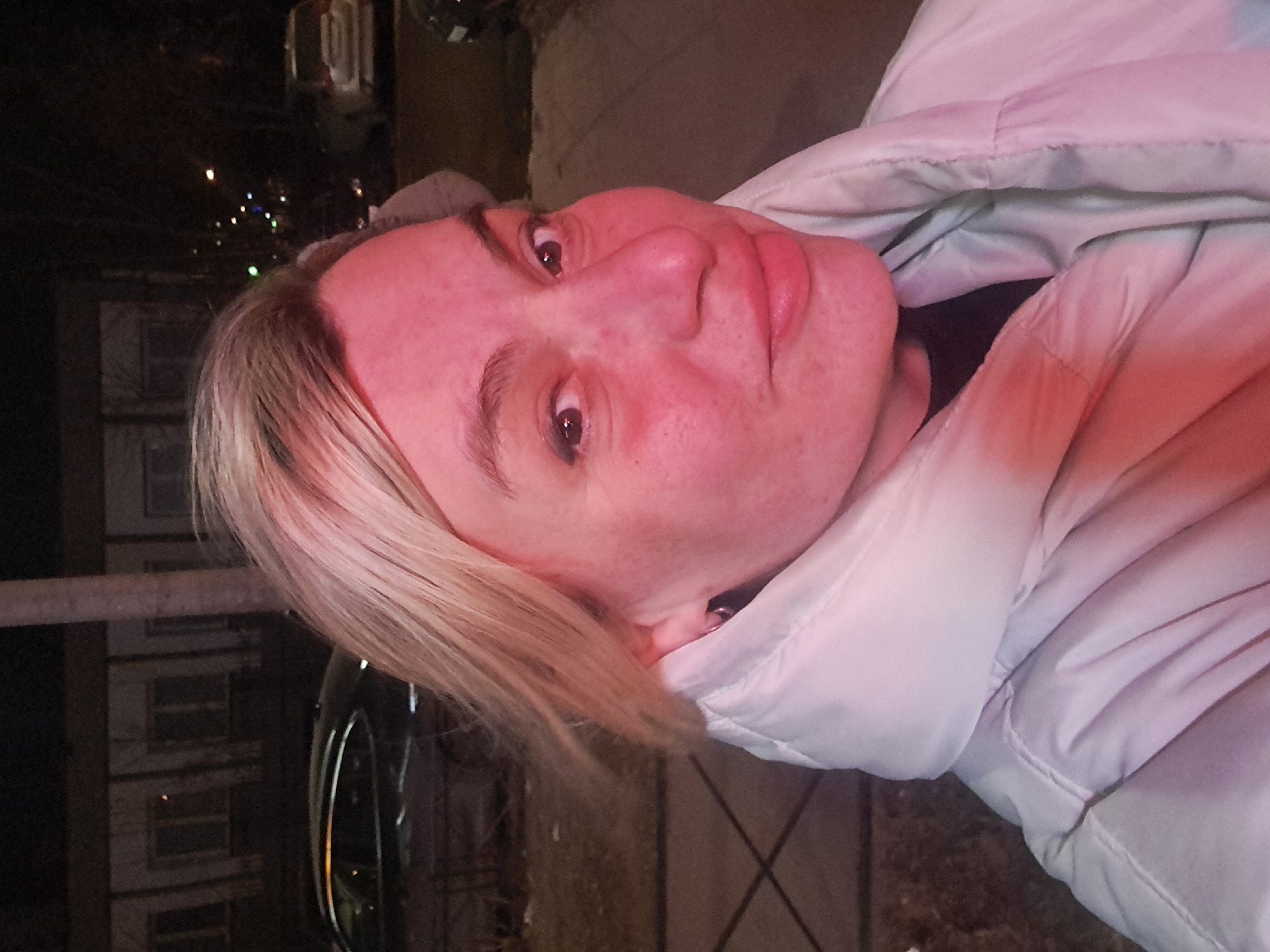 The image depicts an adult female subject in a close-up, selfie-style portrait taken outdoors at night in the city of Bel'kovo, Russia. The subject, with blonde hair, dark eyes, and a neutral facial expression with a slight smirk, is positioned centrally in the frame, facing the camera directly. She is wearing a light-colored (possibly white or light grey) puffer jacket with a high collar, over a dark inner garment. A small silver hoop earring is visible in her left ear. Her face and upper torso are illuminated by a strong reddish-pink light source, causing her skin to appear flushed and pink.
The background is dark and blurred, indicative of nighttime conditions and shallow depth of field. Visible elements include:
- A dark building with multiple illuminated windows in the far left background, suggesting a residential or commercial structure.
- Several parked vehicles, including a dark-colored sedan on the left and a light-colored SUV further back on the right.
- A dark, paved surface, likely a sidewalk or street, extends into the background.
- Bare tree branches are visible against the dark sky.
- Distant streetlights appear as blurred points of light.
- A vertical pole or tree trunk is partially visible behind the subject's head.