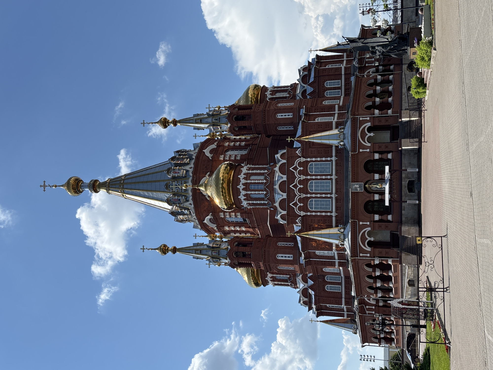 The image shows the Holy Trinity Cathedral in Izhevsk, Russia, on a clear day with scattered clouds. The cathedral is a large, red brick building with multiple golden domes and silver spires topped with crosses. The architectural style is ornate, featuring white decorative elements and numerous arched windows. In front of the cathedral is a paved plaza. To the right of the plaza, there is a bronze statue of two figures holding a flag. The area is bordered by a decorative metal fence and some greenery. There are no people visible in the image.