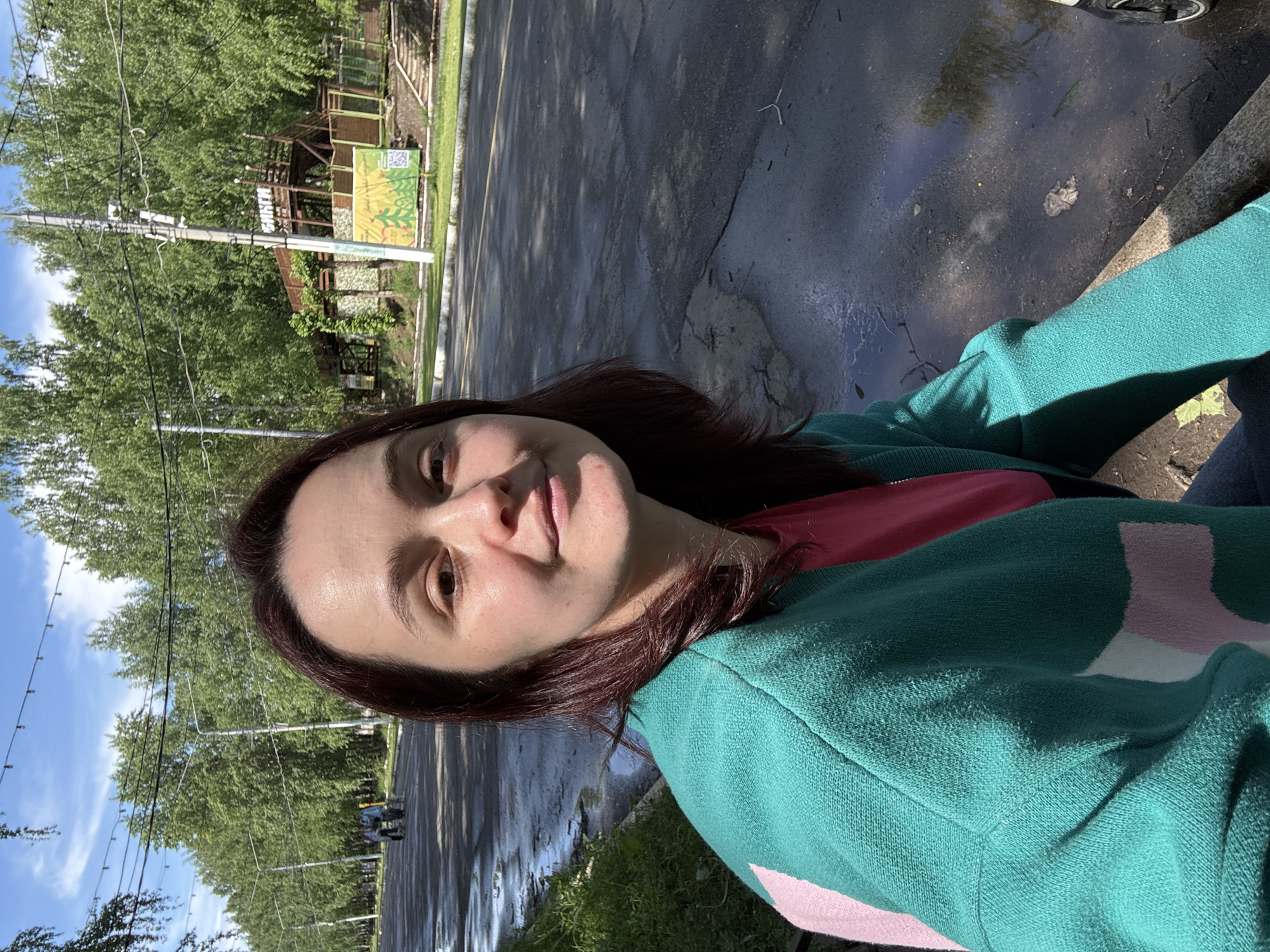 A woman with brown hair and a green sweater is taking a selfie outdoors. The background shows a street, trees, and buildings under a partly cloudy sky. A sign is visible in the background. Based on the style of the building structures, signs, and overall environment, this location is likely Izhevsk, Russia.
