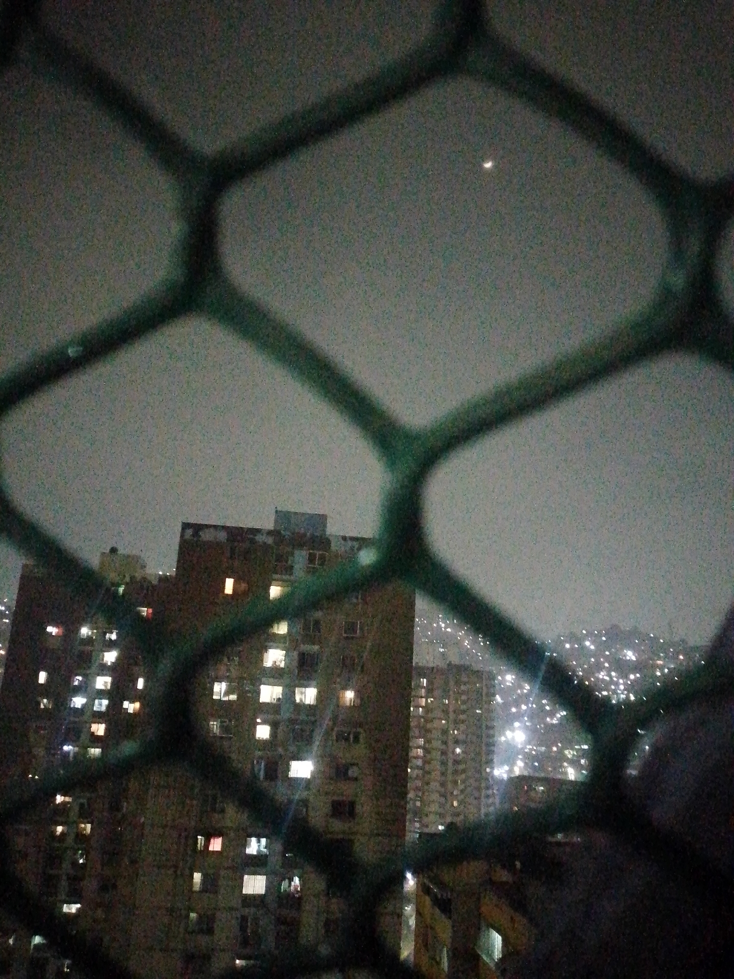 View from a window, through a security grille, showing an overcast night in Caracas, Venezuela. In the center is a tall apartment building with many lit windows. In the distance, a hillside is visible, covered with many lights. A sliver of the moon is in the sky.
