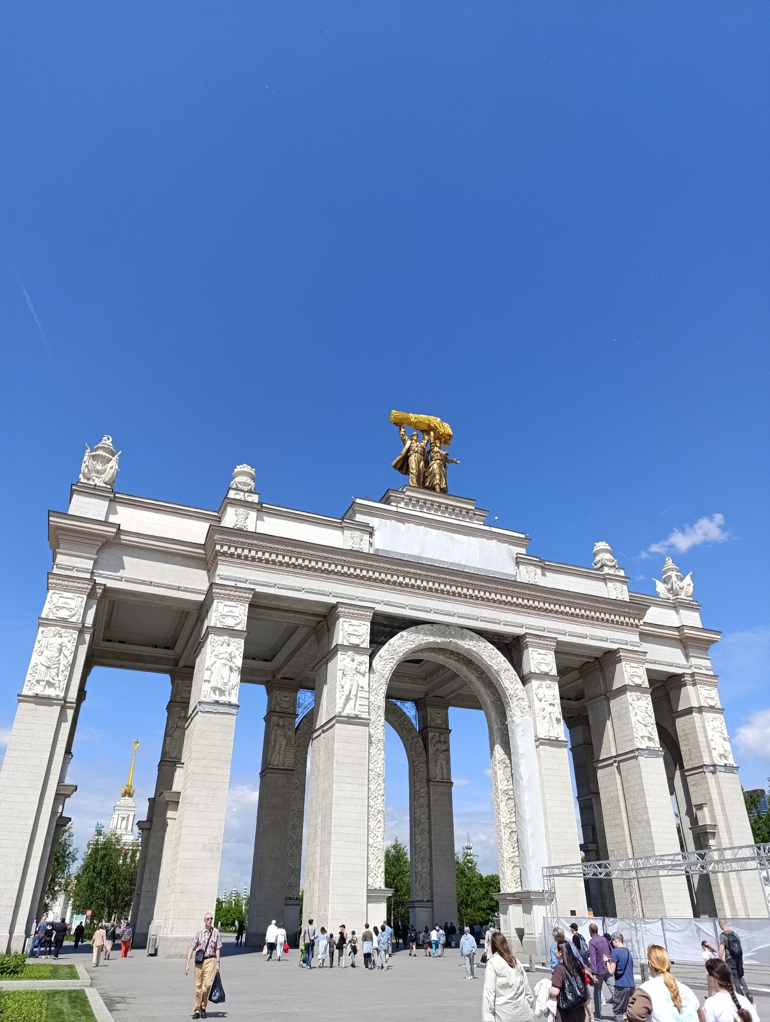 A wide-angle, low-angle shot captures the grand, monumental main entrance archway of what appears to be a large exhibition complex or park. The structure is predominantly white or light-colored stone, featuring multiple colossal columns, ornate arches, and intricate relief sculptures along its facades and within the archways. Each column is adorned with detailed carvings, and decorative crest-like elements punctuate the roofline.
Dominating the top of the central arch is a striking golden sculpture depicting two figures, one male and one female, holding a massive bundle of wheat or grain above their heads. This iconic artwork is brightly illuminated by the sun.
Below and around the archway, numerous people are scattered across wide paved areas, casually walking and exploring. They are dressed in a variety of light, casual clothing, suggesting warm, sunny weather. To the left, in the background, a distinctive golden-spired building with a white base is visible, partially framed by green trees. On the right side, temporary metal scaffolding or event staging structures are visible, hinting at ongoing activities or preparations.
The scene is bathed in bright daylight under a clear, deep blue sky with only a few faint clouds. The strong sunlight casts clear shadows, indicating it is likely midday or early afternoon. The ground consists of paved walkways and neatly manicured green lawns. The grand architecture, particularly the golden sculpture and the distant spire, are strong indicators of the VDNKh (Exhibition of Achievements of National Economy) in Moscow, Russia. No discernible text is visible in the image.