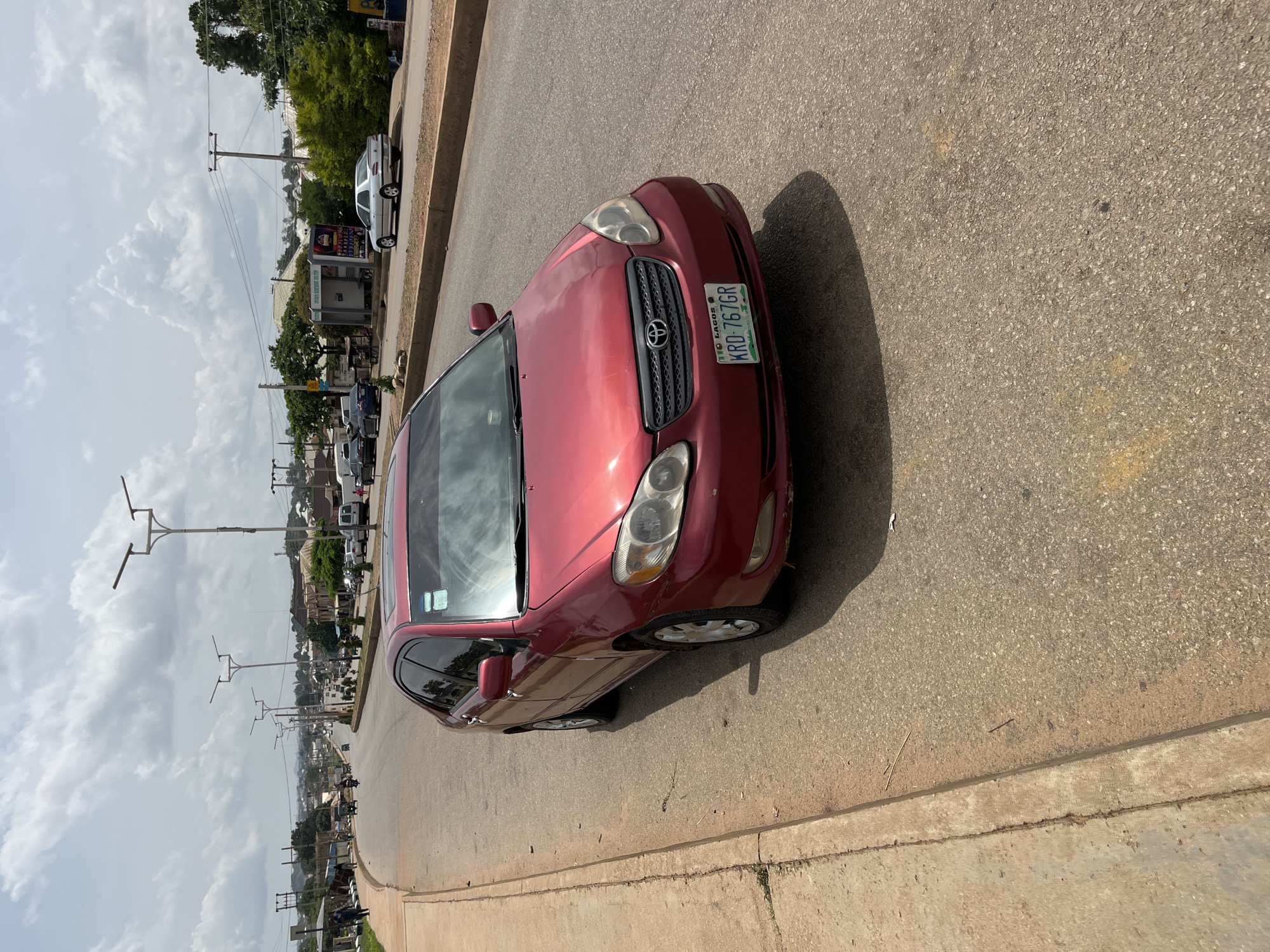 A red sedan, identified by the license plate KRD-767GR, is parked on the right side of a paved road. The car faces the viewer, with its front slightly angled towards the left. The road is lined with a concrete sidewalk on the left, and the right side of the road has a curb. Beyond the curb, there are green trees and buildings. In the background, utility poles with wires stretch across the sky. The sky is partly cloudy. The scene appears to be in Bukuru, Nigeria.