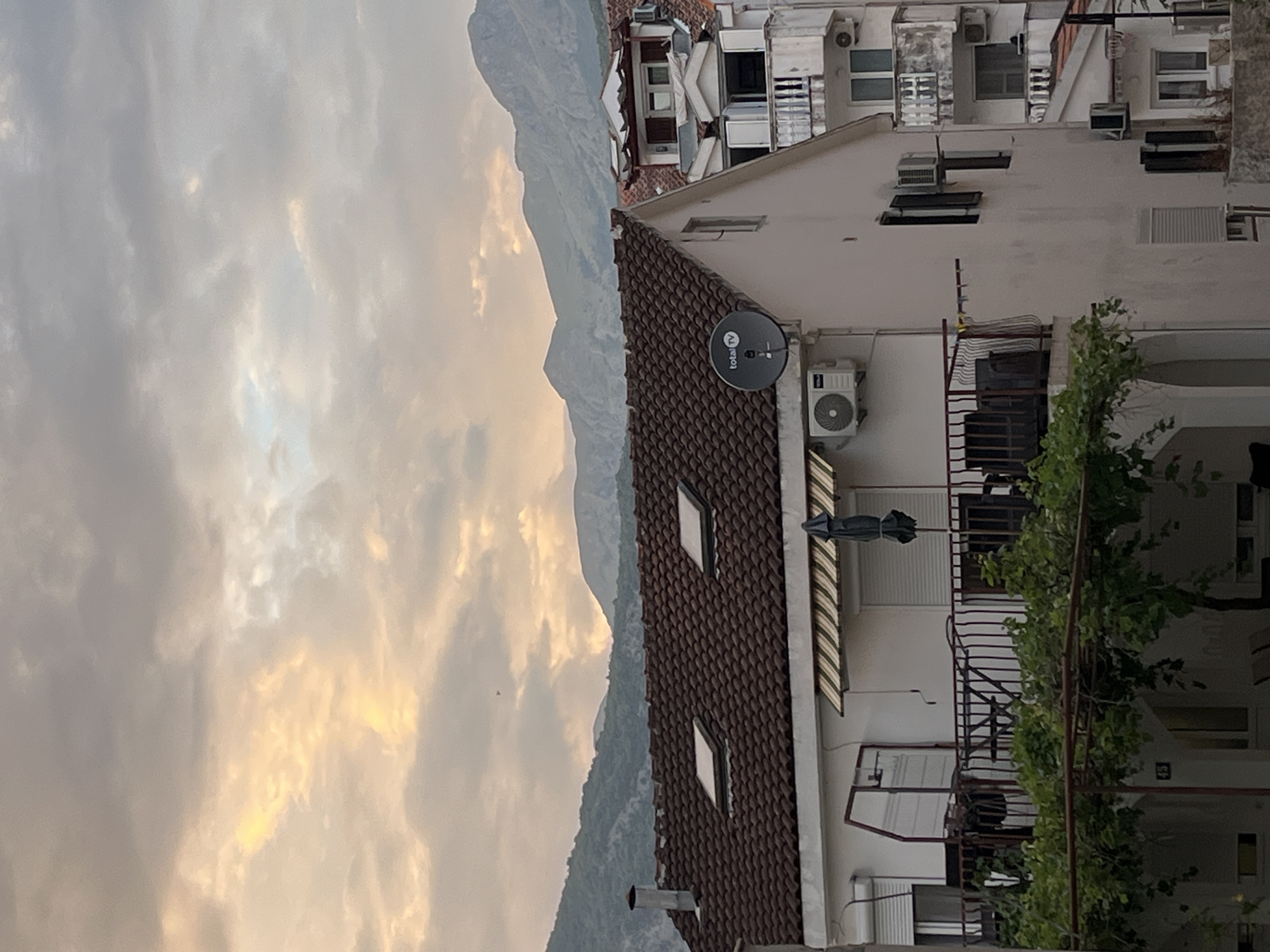 The image shows a view of buildings and mountains in Budva, Montenegro. The foreground features a white building with a red tile roof and two skylights. A balcony with a dark metal railing is visible on the second floor, along with a patio table and chairs, and an umbrella. An air conditioning unit is mounted on the wall. A satellite dish is also attached to the side of the building. To the right, another building with white balconies and windows is visible. In the background, lush green mountains rise against a cloudy sky with warm, soft light filtering through. The overall scene depicts a residential area within a mountainous coastal landscape.