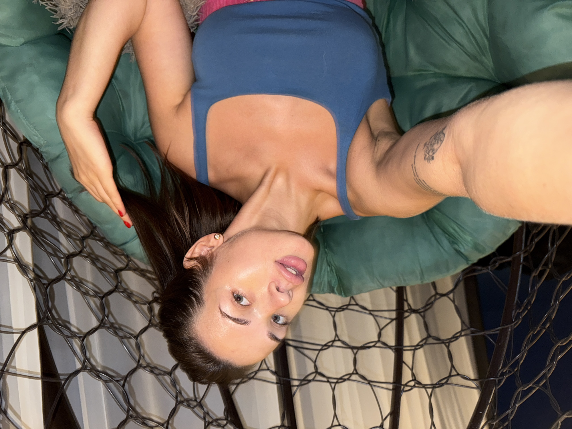 A woman is sitting in a green chair. She is wearing a blue tank top and pink shorts. She has long brown hair and is wearing makeup. She is holding a phone in her right hand and taking a selfie. The chair is in front of a window with a black mesh curtain. The woman appears to be in her home in Lobanovo, Russia.