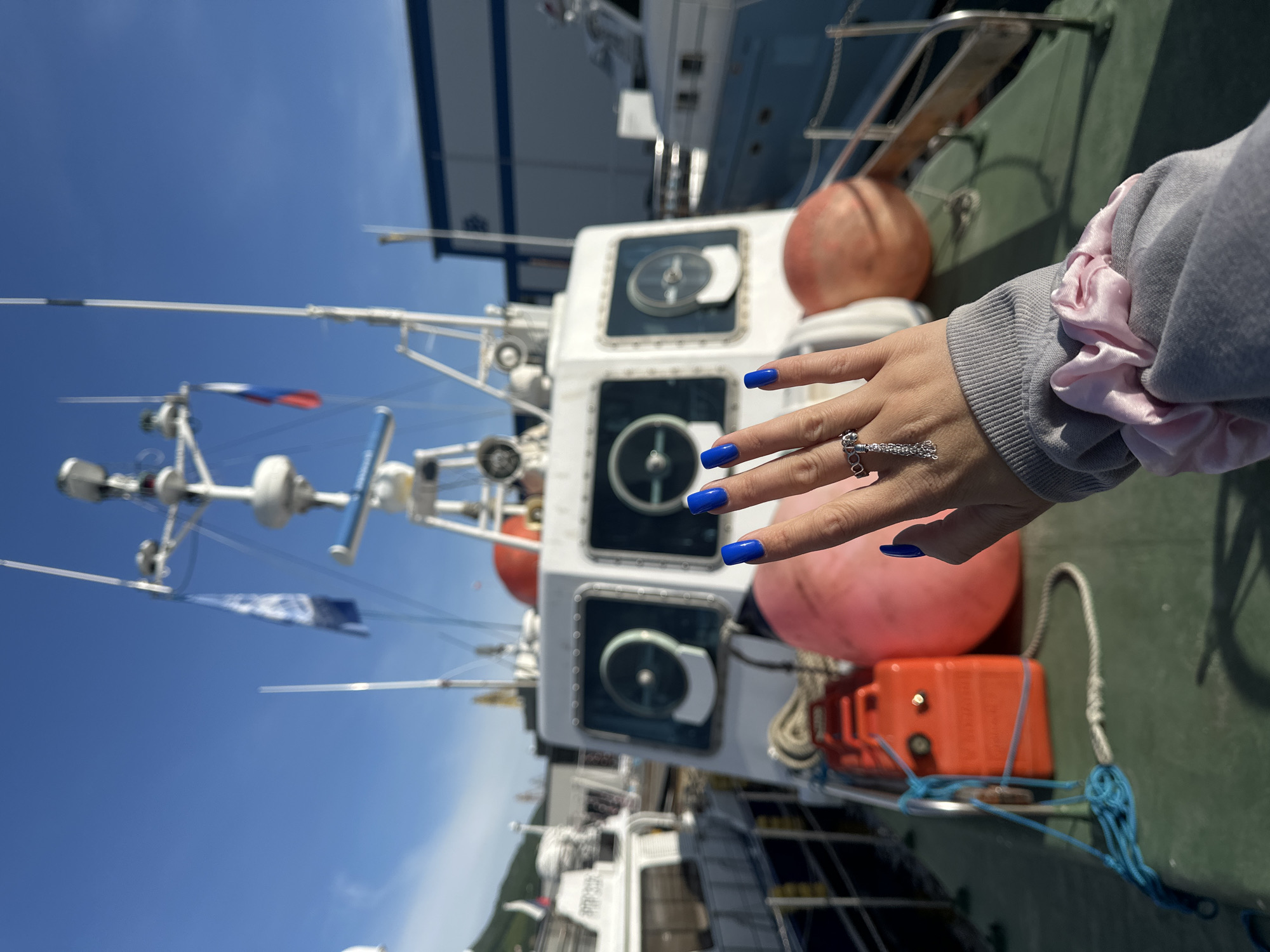 The image captures a hand with bright blue fingernails and a silver ring with a dangling chain and small stones, extending towards the viewer. The hand is positioned over a grey sweatshirt with a pink scrunchie on the wrist. In the background, a white boat with several windows and nautical equipment, including flags, is docked. The boat is adorned with large orange buoys. To the right of the boat, a building with blue and white accents is visible. The scene appears to be a harbor, possibly in Petropavlovsk-Kamchatsky, Russia, given the presence of a Russian flag on the boat. The sky is clear and blue, indicating fair weather. The overall impression is one of a casual moment captured by the water, with the boat serving as a prominent backdrop.